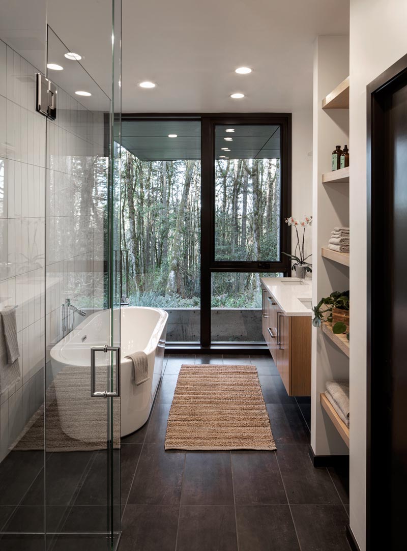 In this modern master bathroom, a freestanding bathtub and the shower are positioned against one wall, while a floating wood vanity with dual sinks and a wall of open shelving are located on the opposite wall. #MasterBathroom #ModernBathroom #BathroomIdeas