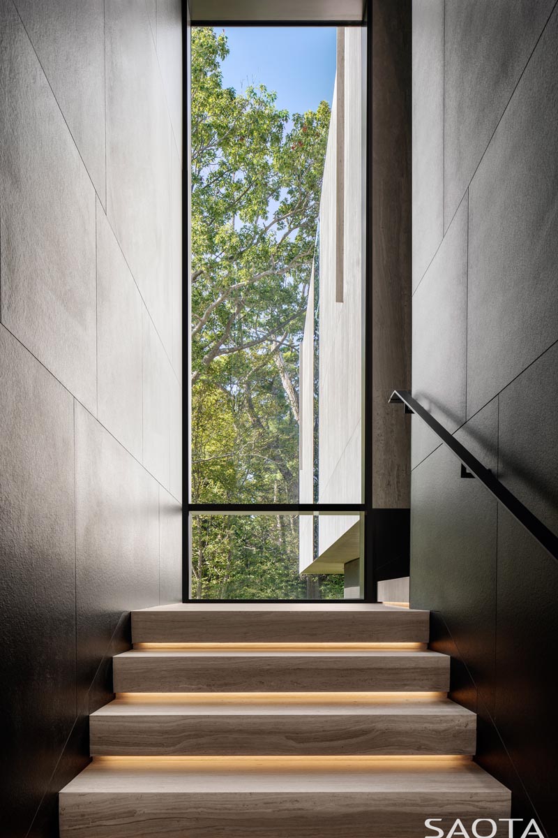 This modern staircase is lined with the same ceramic panels as the exterior of the house, while the wood stair treads have hidden lighting underneath them, and a tall window provides views of the trees. #StairDesign #TallWindow #StairsWithLighting