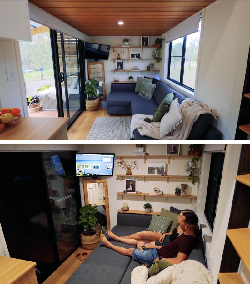 Stepping through the sliding doors of this modern tiny house, you're immediately in the living room that features a wall of wood bookshelves, and a regular size couch with hidden storage and a pull-out section that transforms it into a comfortable place to watch tv. #TinyHouse #TInyHouseLivingRoom #SmallLiving