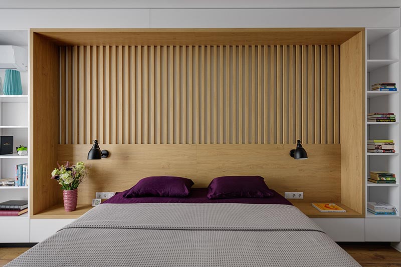 The design of this custom wall in a modern bedroom, combines wood and white details, and integrates the bed frame, a headboard, side tables, and bookshelves. #BedroomDesign #ModernBedroom #Headboard #Bookshelves