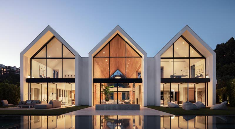 The eye-catching gabled design of this modern barn-inspired house, is easily seen upon approach to the house, and once inside, the wall of windows and doors at the other end of the wood-lined great room showcase the swimming pool and view. #ModernHouse #ModernArchitecture #HouseDesign #GabledRoof #GreatRoom