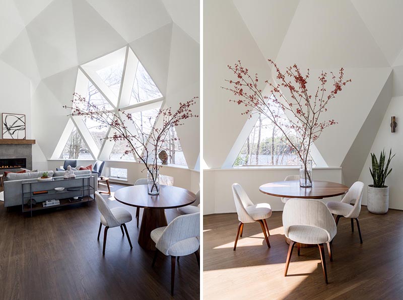 The newly updated interior of this geodesic dome house features bright white walls and modern furnishings feature throughout, like in the living room. The fireplace, which sits next to a large section of windows, has a limestone surround topped with a custom oak mantel that complements the hardwood flooring. #GeodesicDome #LivingRoom #Fireplace #TriangleWindows #CasualDining