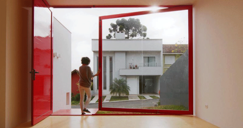 When the front door of this modern house is open, the large pivoting window can be moved by pushing the frame to open it up completely, exposing the interior of the home to the small grass patio. and front yard. #PivotingWindow #RedFrontDoor #WindowIdeas