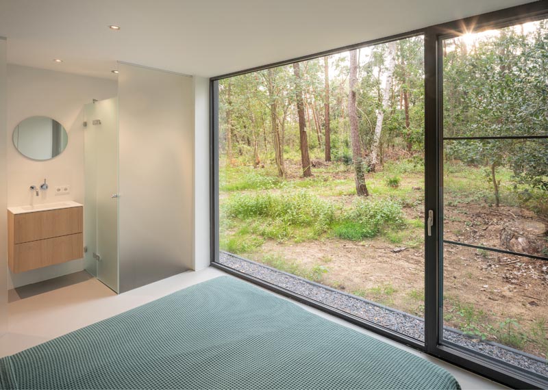 In this modern master bathroom, there's a large floor-to-ceiling picture window, as well as a bathroom that has a shower surrounded by a frosted screen. #FrostedShowerScreen #MasterBedroom #PictureWindow