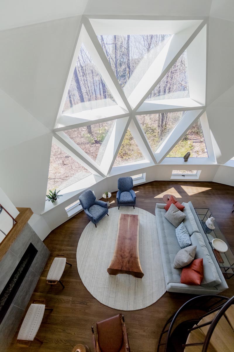 The newly updated interior of this geodesic dome house features bright white walls and modern furnishings feature throughout, like in the living room. The fireplace, which sits next to a large section of windows, has a limestone surround topped with a custom oak mantel that complements the hardwood flooring. #GeodesicDome #LivingRoom #Fireplace #TriangleWindows #InteriorDesign