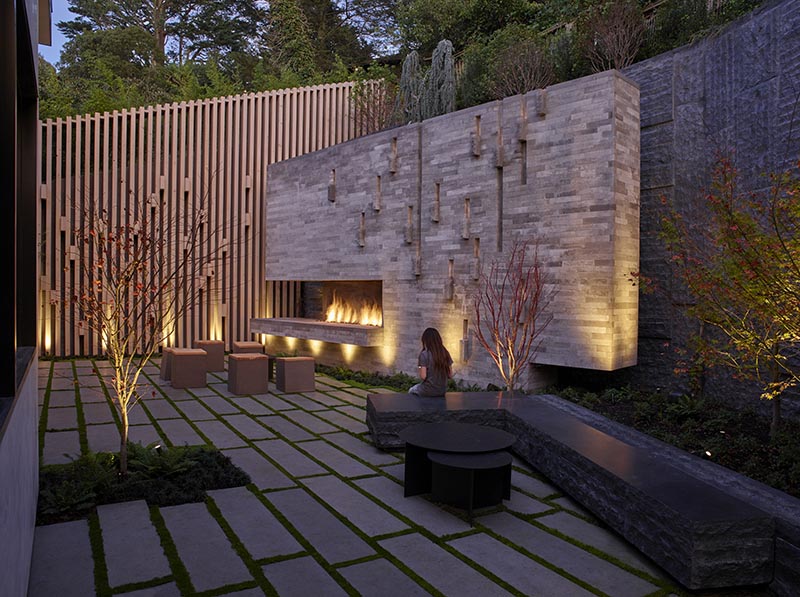 This modern courtyard has Basalt-clad retaining walls that hold back the hillside and frame the courtyard, while the limestone hearth anchors the garden, and a panel of blackened steel creates a backdrop for a built-in barbeque. #LandscapeDesign #Landscaping #Courtyard #RetainingWalls #OutdoorFireplace