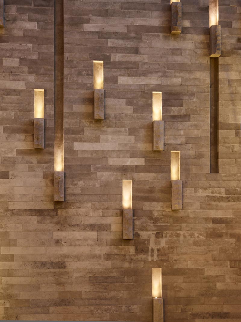 Notches on the face of an outdoor feature wall create channels of light that are punctuated by stone accents, creating a sculptural lantern-like light feature at night. #OutdoorLighting #LandscapeDesign #Landscaping