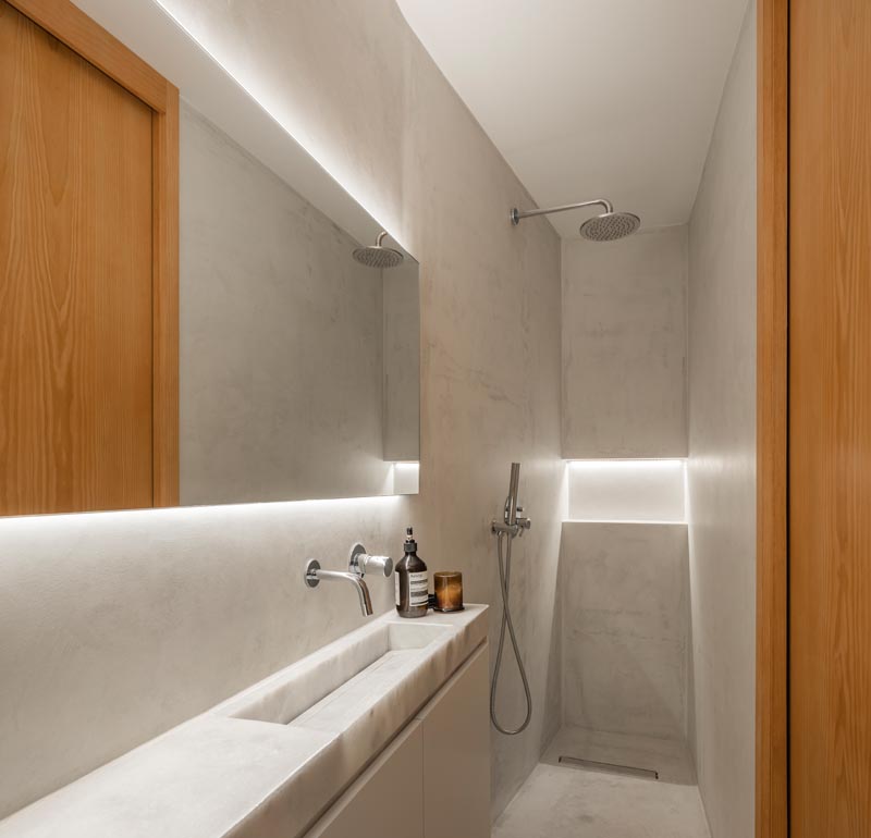 This small and narrow modern bathroom features a long thin vanity along one wall, with the shower at one end and the toilet at the other. Hidden lighting behind the mirror and in the shower niche help to keep the space bright. #SmallBathroom #NarrowBathroom #BathroomDesign #HiddenBathroomLighting
