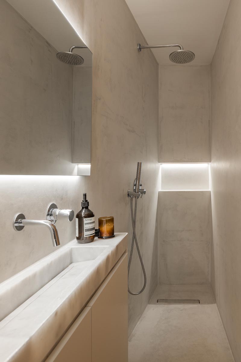 This small and narrow modern bathroom features a long thin vanity along one wall, with the shower at one end and the toilet at the other. Hidden lighting behind the mirror and in the shower niche help to keep the space bright. #SmallBathroom #NarrowBathroom #BathroomDesign #HiddenBathroomLighting #InvisibleDrain