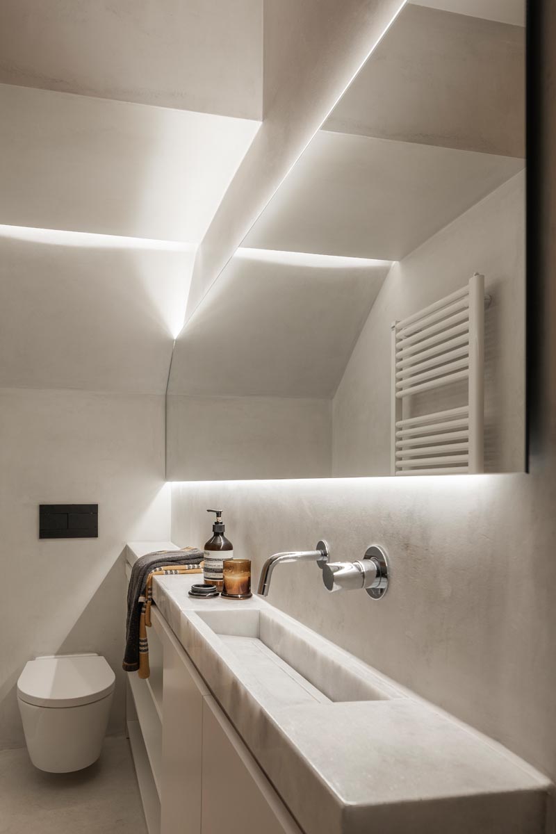 This small and narrow modern bathroom features a long thin vanity along one wall, with the shower at one end and the toilet at the other. Hidden lighting behind the mirror and in the shower niche help to keep the space bright. #SmallBathroom #NarrowBathroom #BathroomDesign #HiddenBathroomLighting