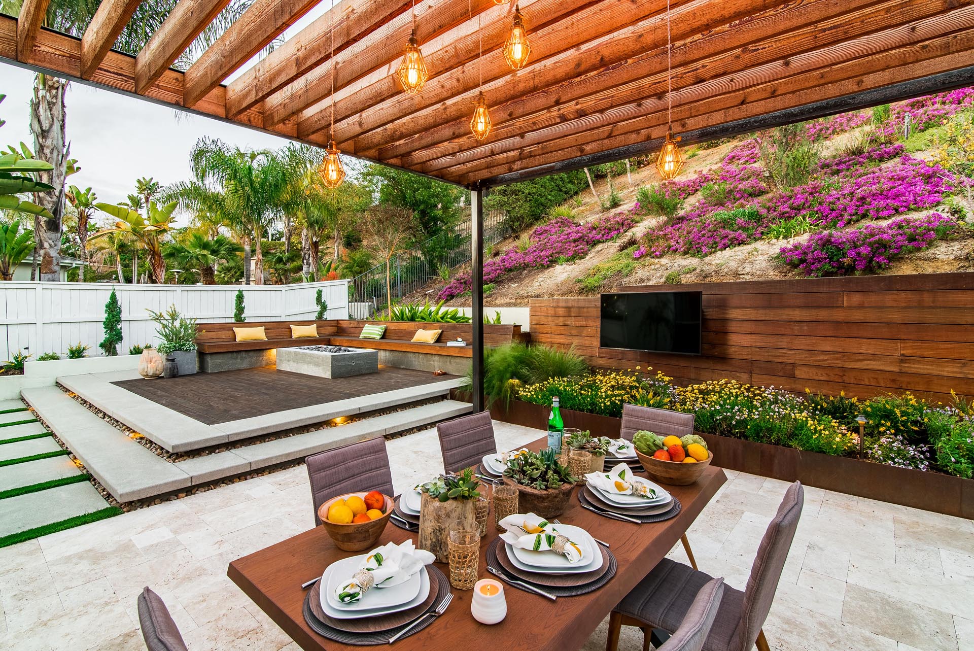 A modern yard with an elevated fire pit, deck, pergola, and outdoor tv.