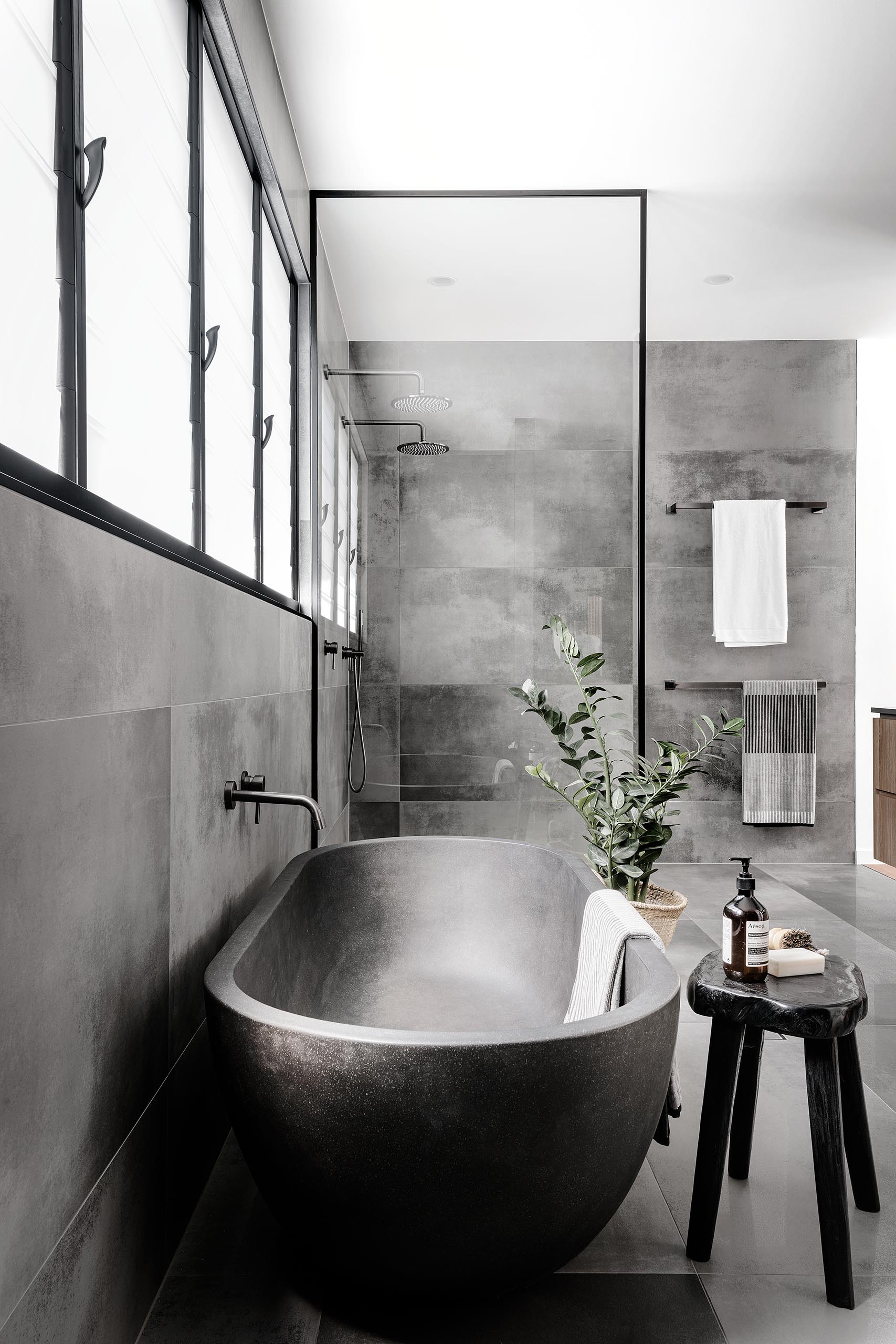 A modern master bathroom with large format grey tiles, a  grey freestanding bathtub, and a black framed glass shower screen.