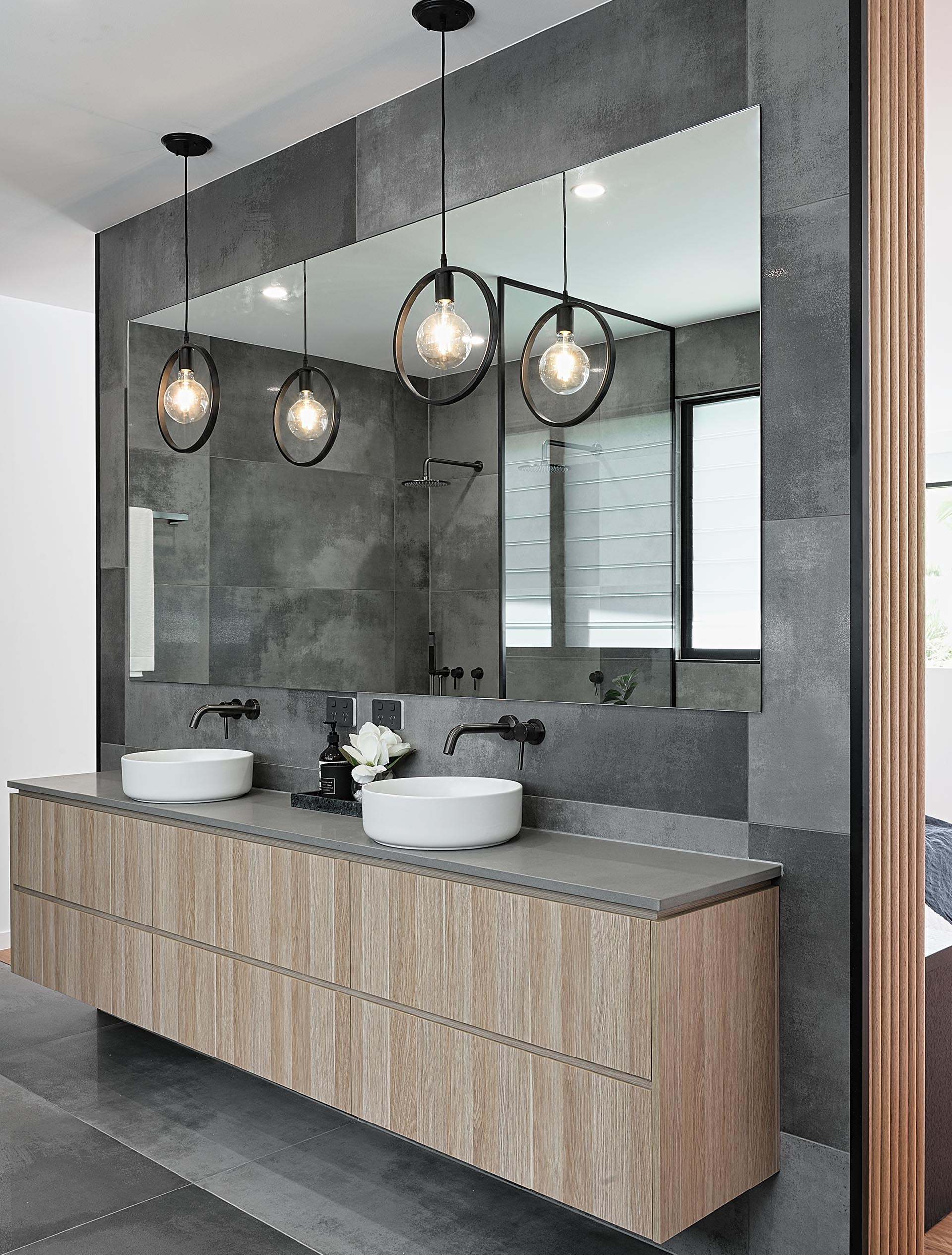 A modern bathroom vanity with wood cabinets, white sinks, a grey countertop, black accents, and two pendant lights.