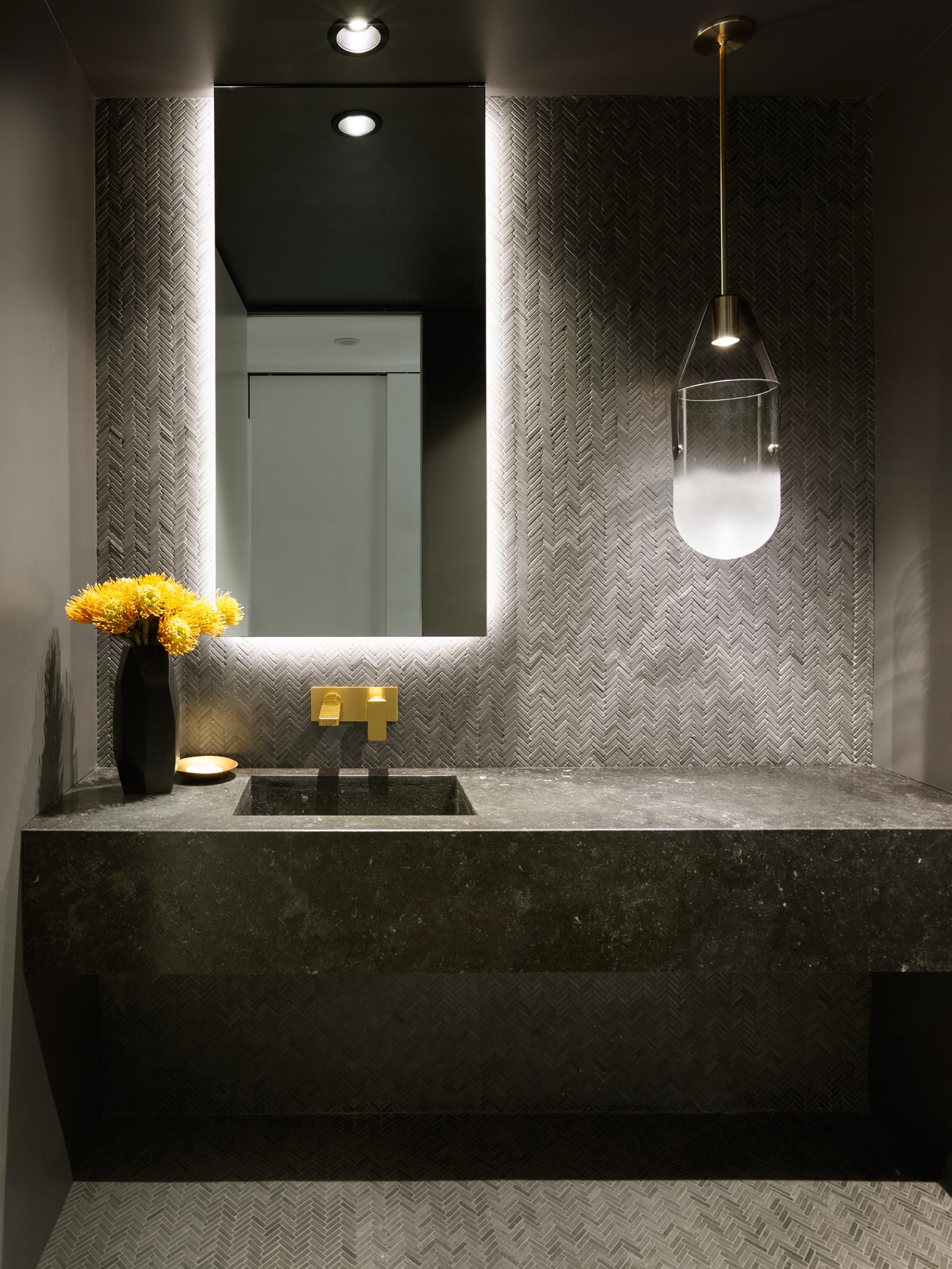 A modern powder room with dark grey tiles, a backlit mirror, and a glass pendant light.