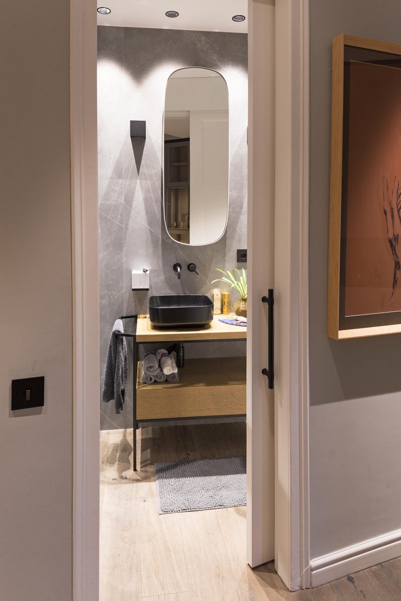 A modern grey bathroom with a pill-shaped mirror, a sliding door, black accents, and a wood vanity.