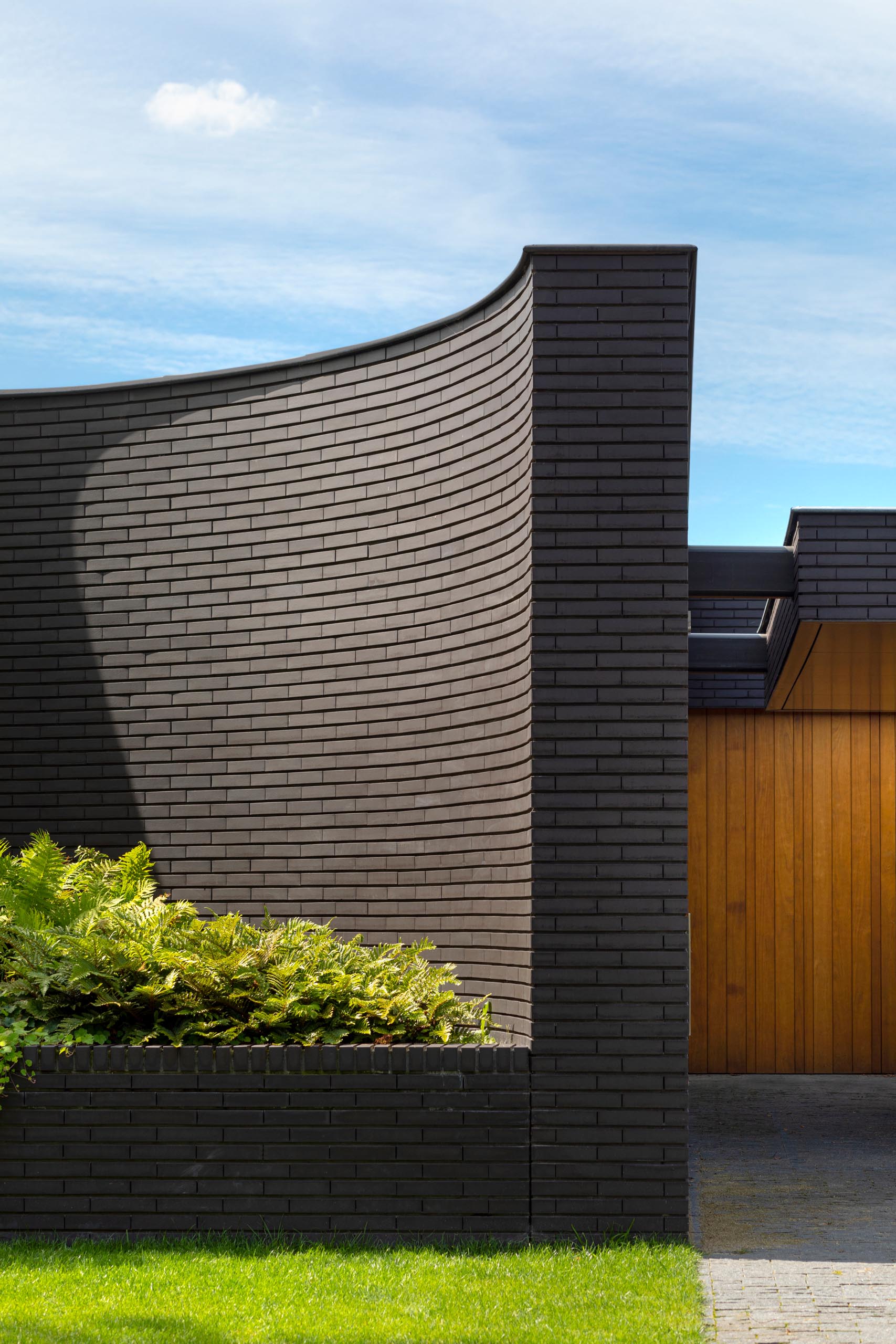 The black brick facade of this modern home features curves and has large integrated planters that highlight the lush greenery.