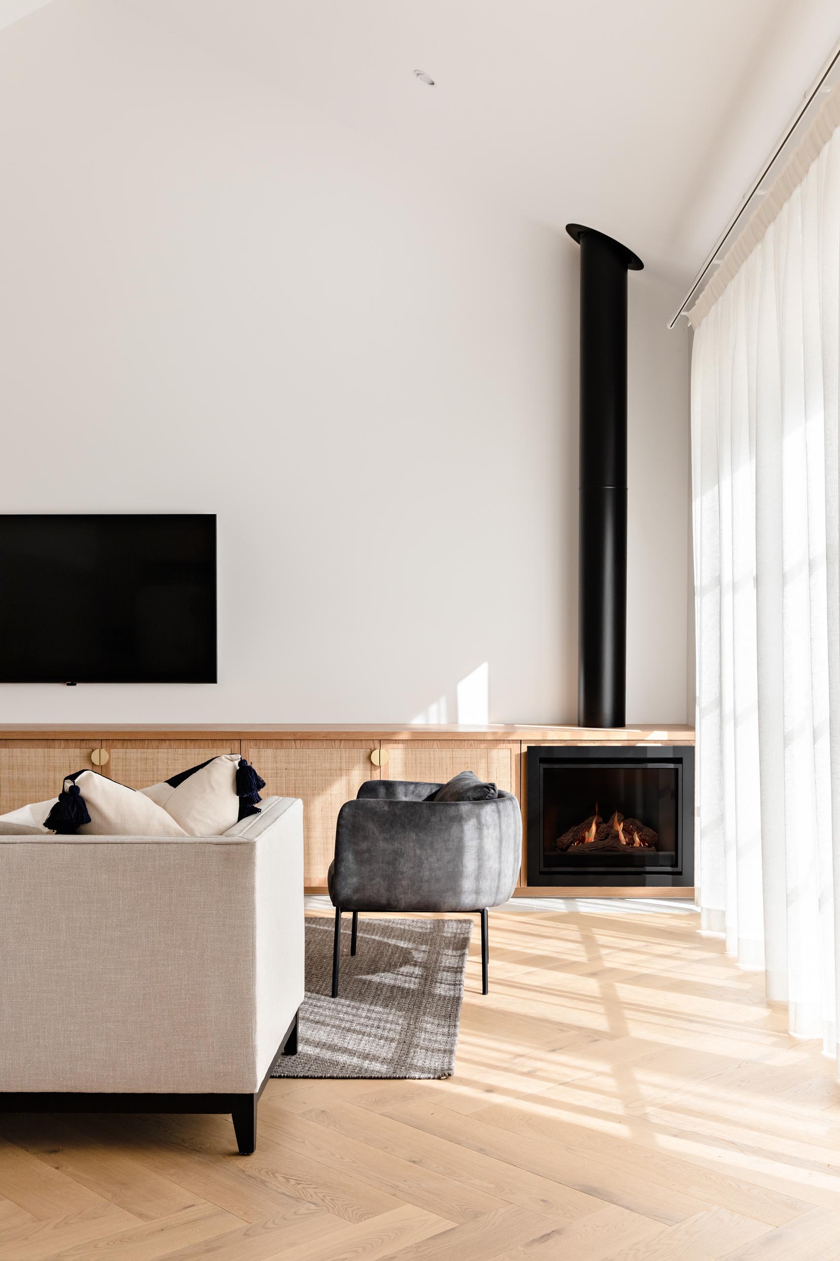 A modern living room with a custom oak and rattan cabinet and a black gas fireplace with exposed flue.