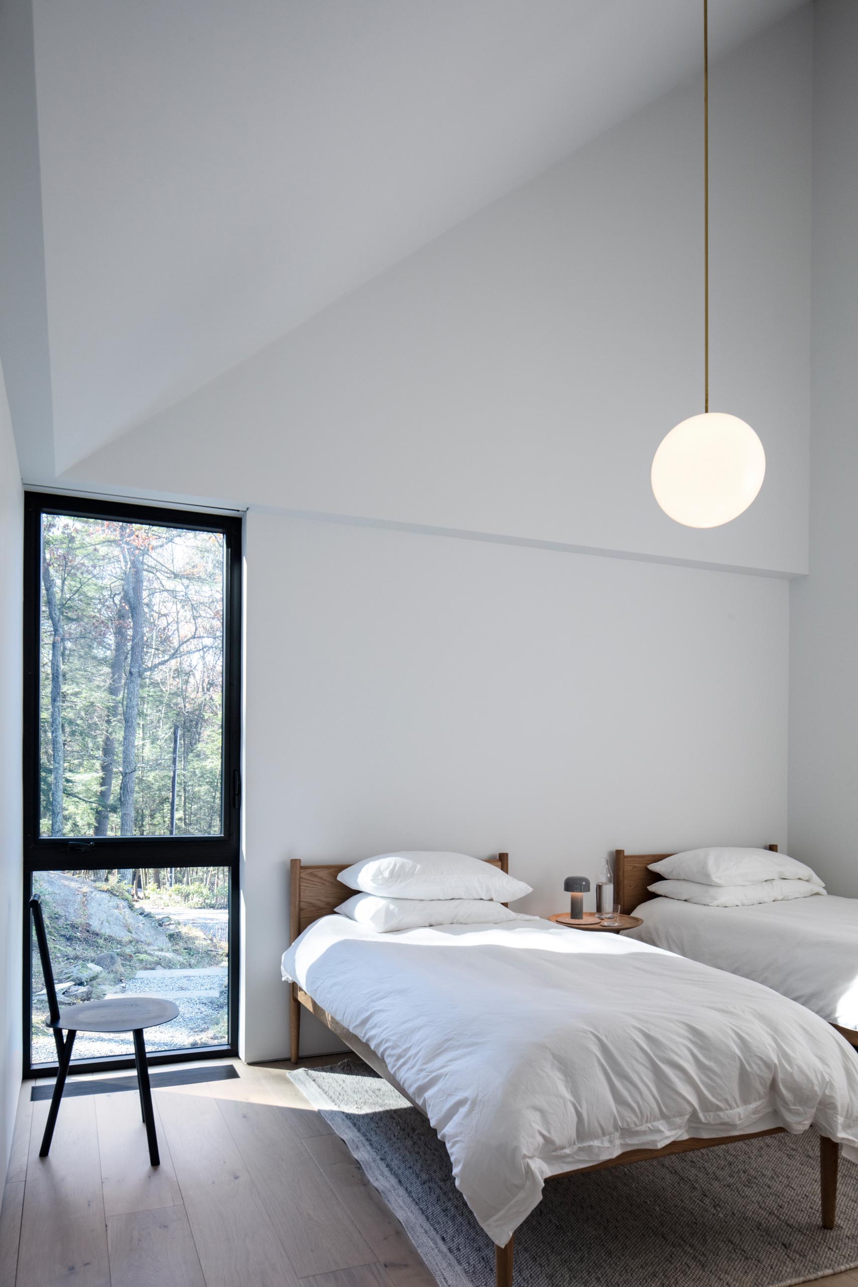 A minimalist guest bedroom with two beds, a modern black chair, white walls, and a single pendant light.