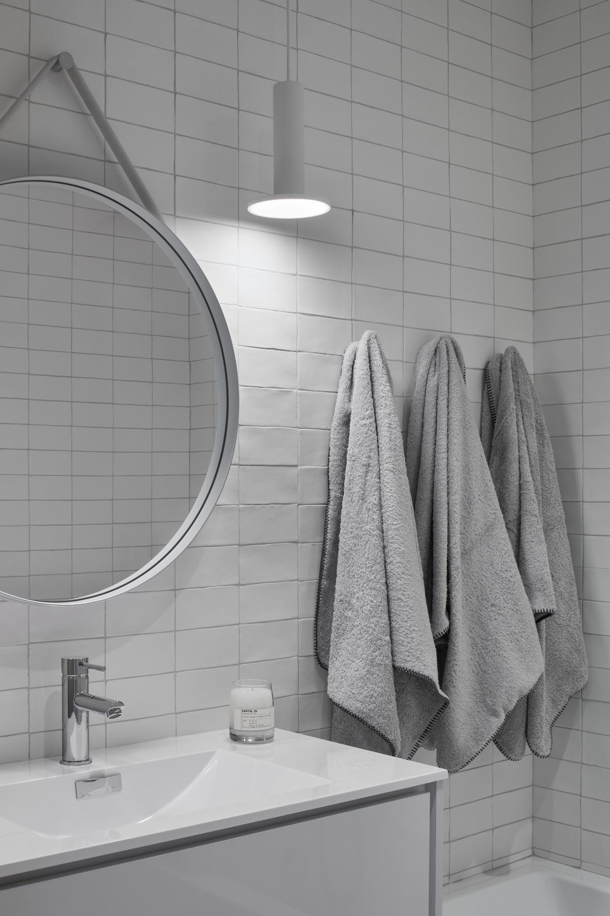 A modern white bathroom with white tiles, white pendant light, white framed round mirror, and white vanity.