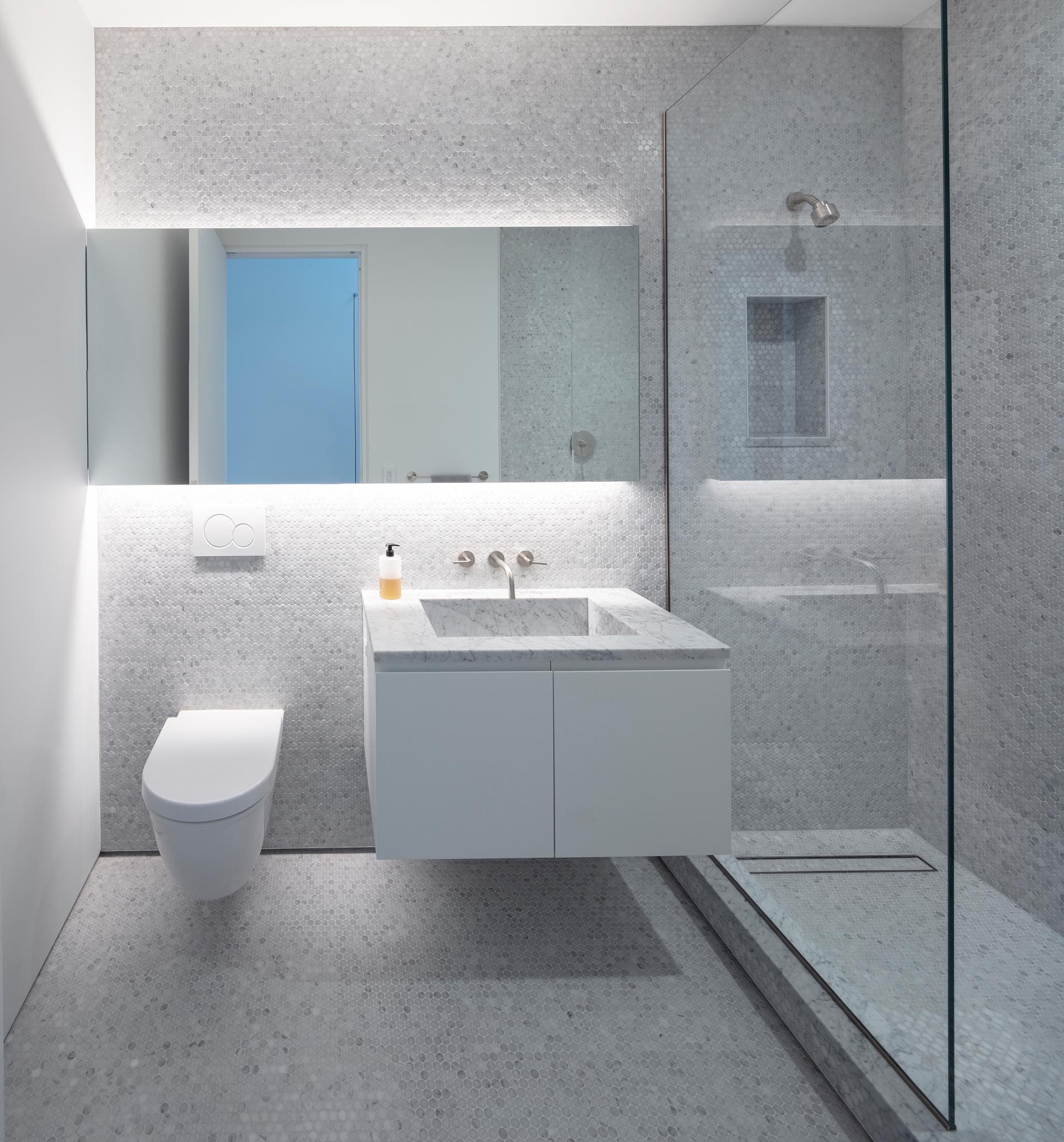 A modern bathroom with floor-to-ceiling grey penny tiles, a glass shower screen, a backlit mirror, and a floating vanity.