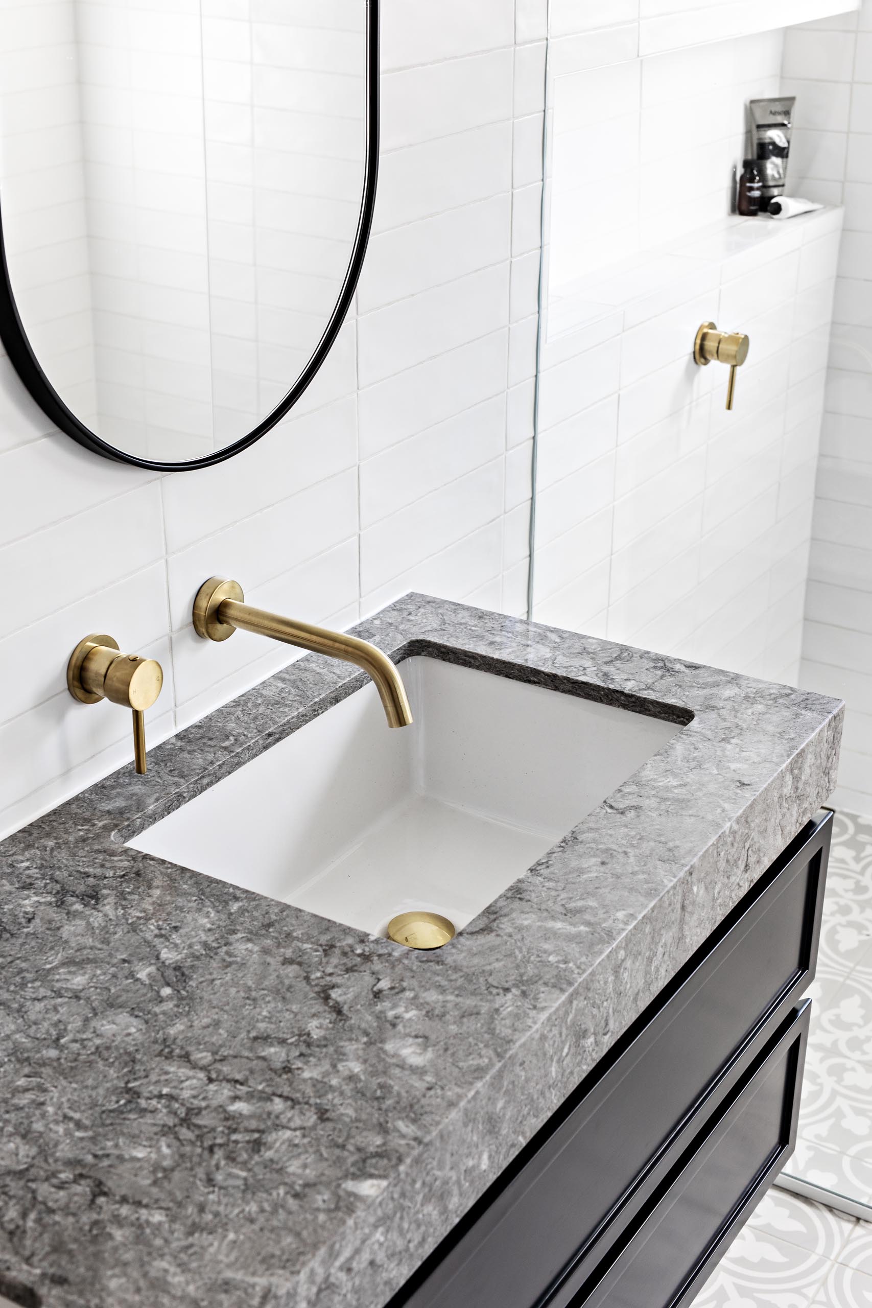 patterned floor tiles, a floating black vanity topped with grey stone, white subway tiles that cover the walls, oval black-framed mirrors, and brass hardware