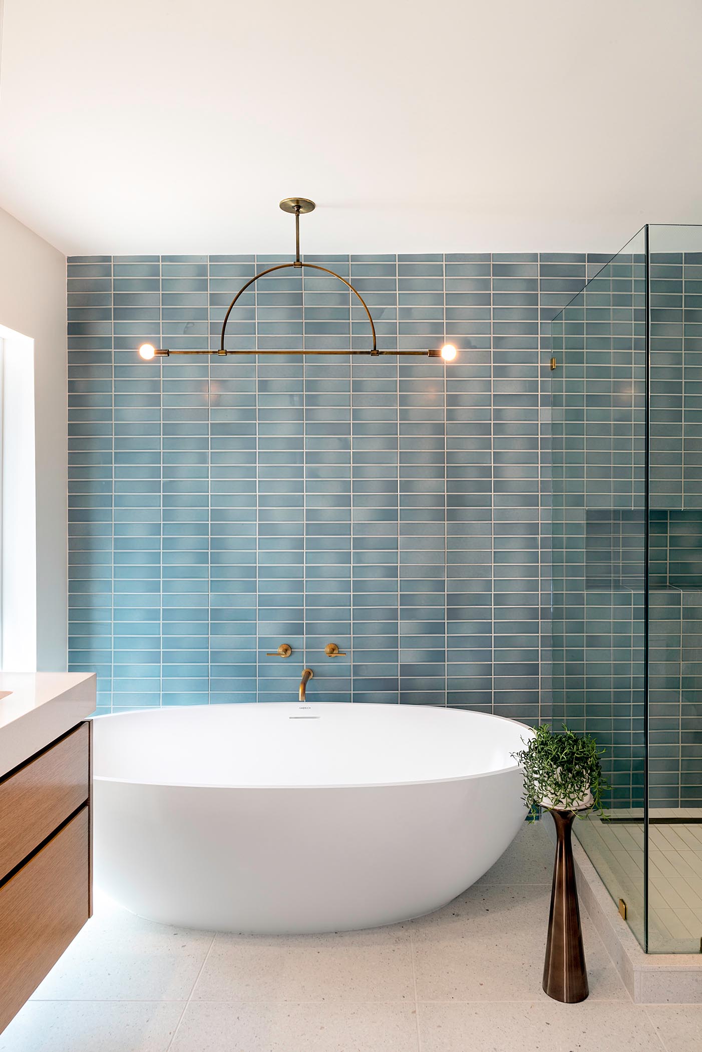 This modern bathroom with blue rectangular wall tile, a minimalist light fixture, a freestanding white bathtub, and brass fixtures.