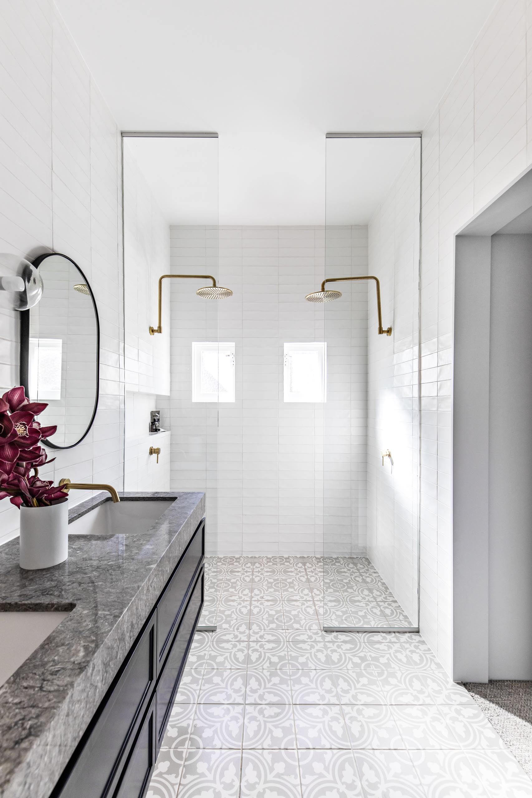 A modern bathroom with patterned floor tiles, a floating black vanity topped with grey stone, white subway tiles that cover the walls, oval black-framed mirrors, brass hardware, and floor-to-ceiling glass shower screens.