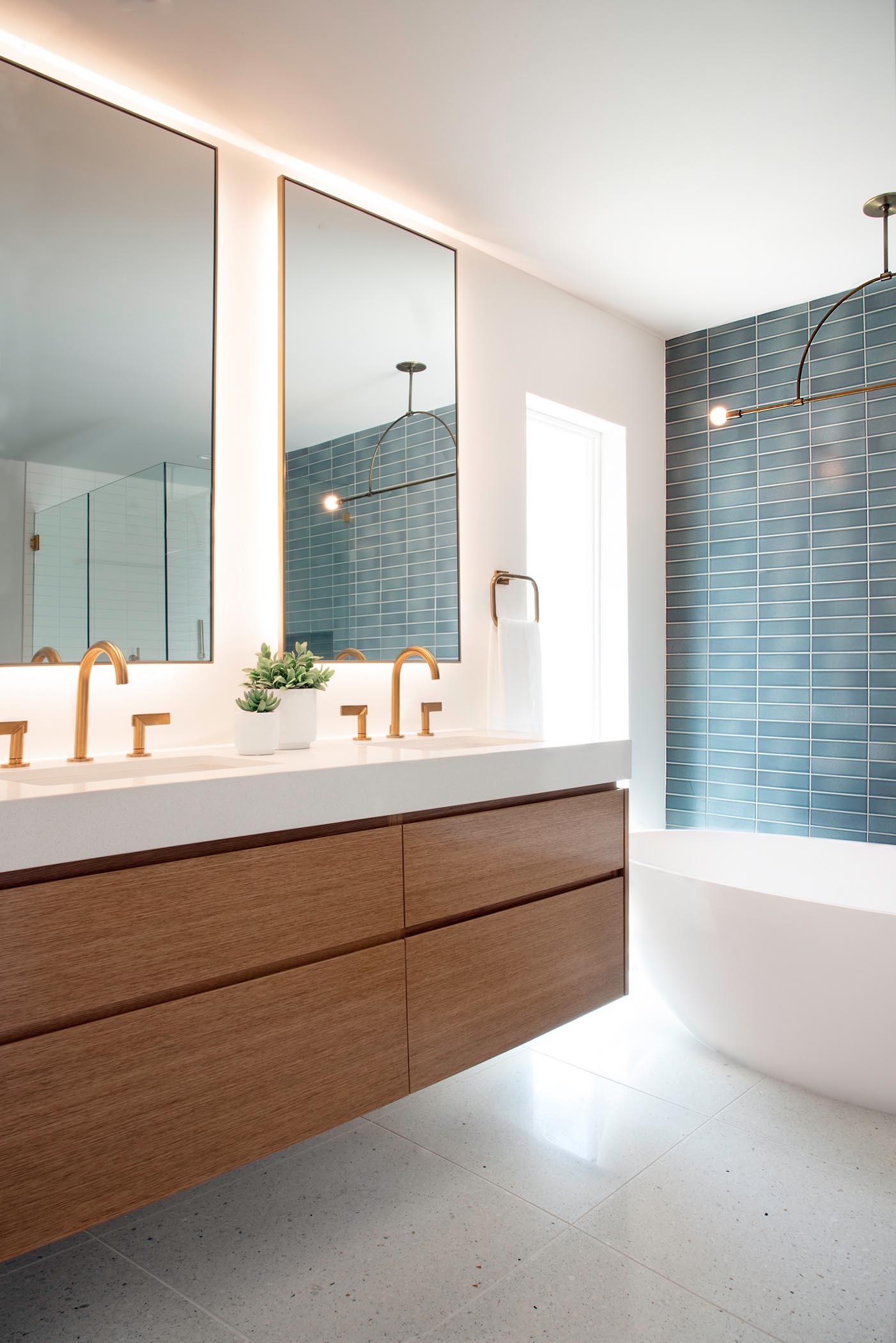 This modern bathroom includes a wall mounted wood vanity, blue and white rectangular wall tile, a minimalist light fixture, custom backlit mirrors, a freestanding white bathtub, and brass fixtures.