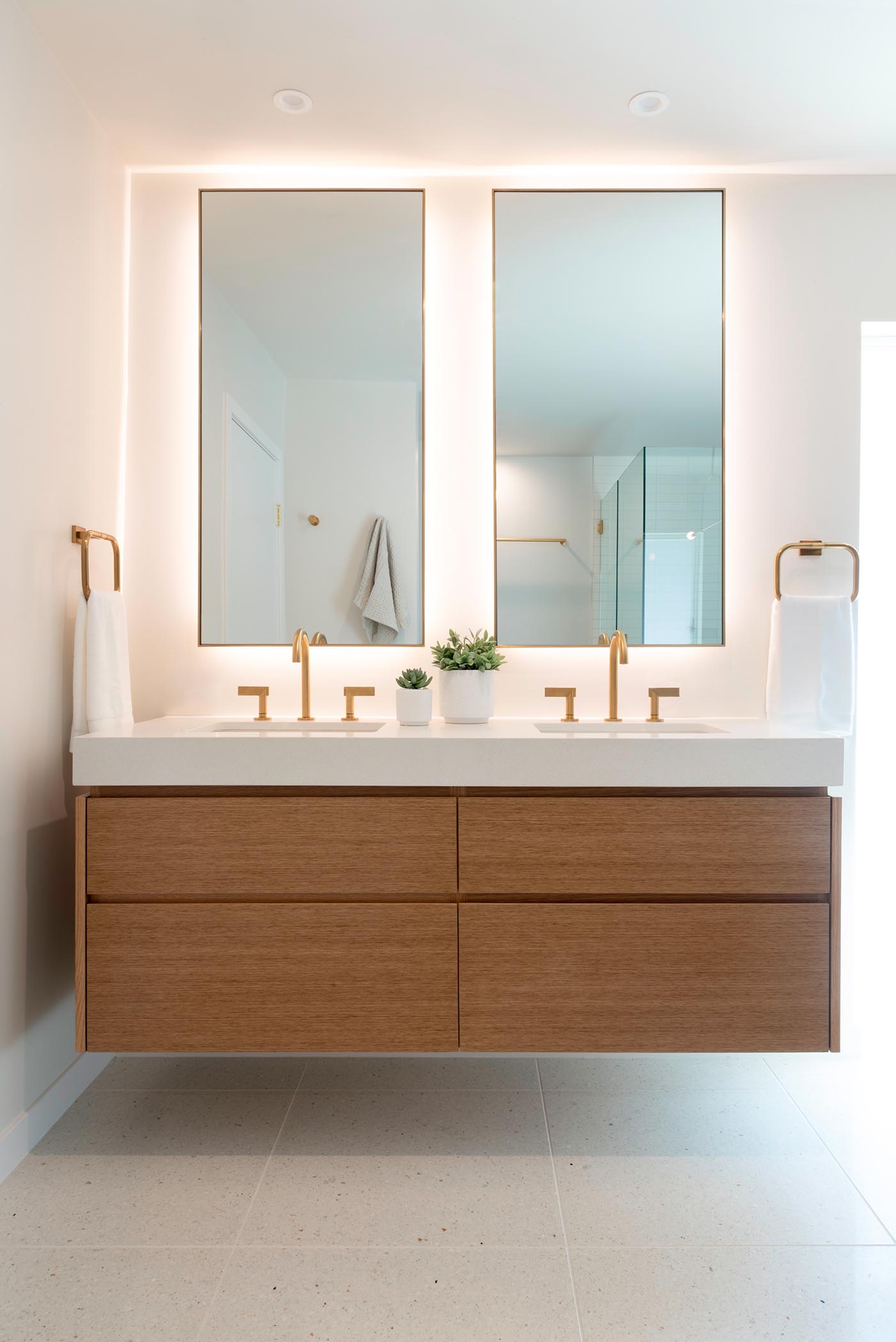 This modern bathroom includes a wall mounted wood vanity with custom backlit mirrors, and brass fixtures.