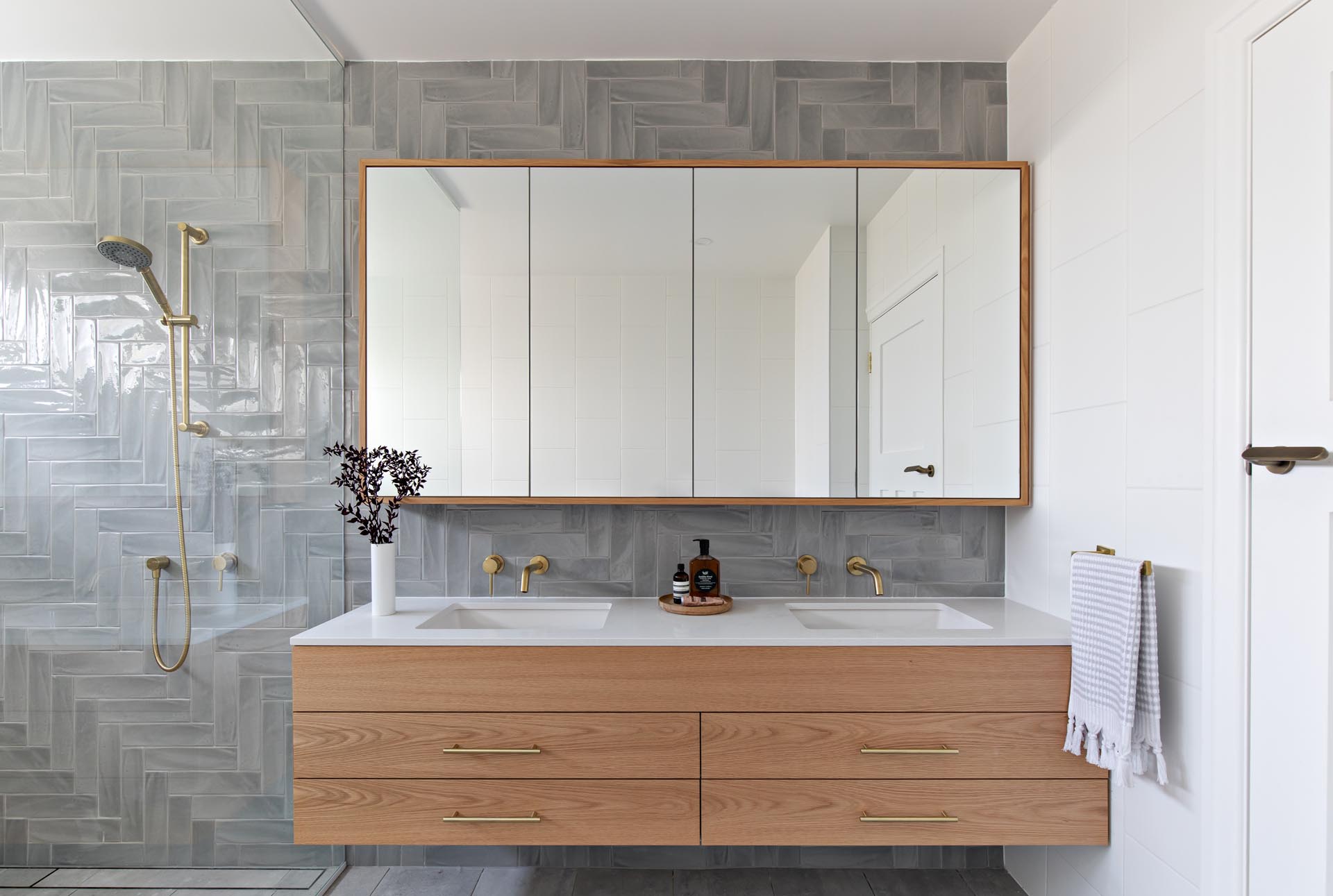 A modern bathroom with grey tiles installed in a herringbone pattern cover the wall, a floating wood vanity with a white countertop, and a glass shower screen that separates the shower and freestanding bathtub from the rest of the room.