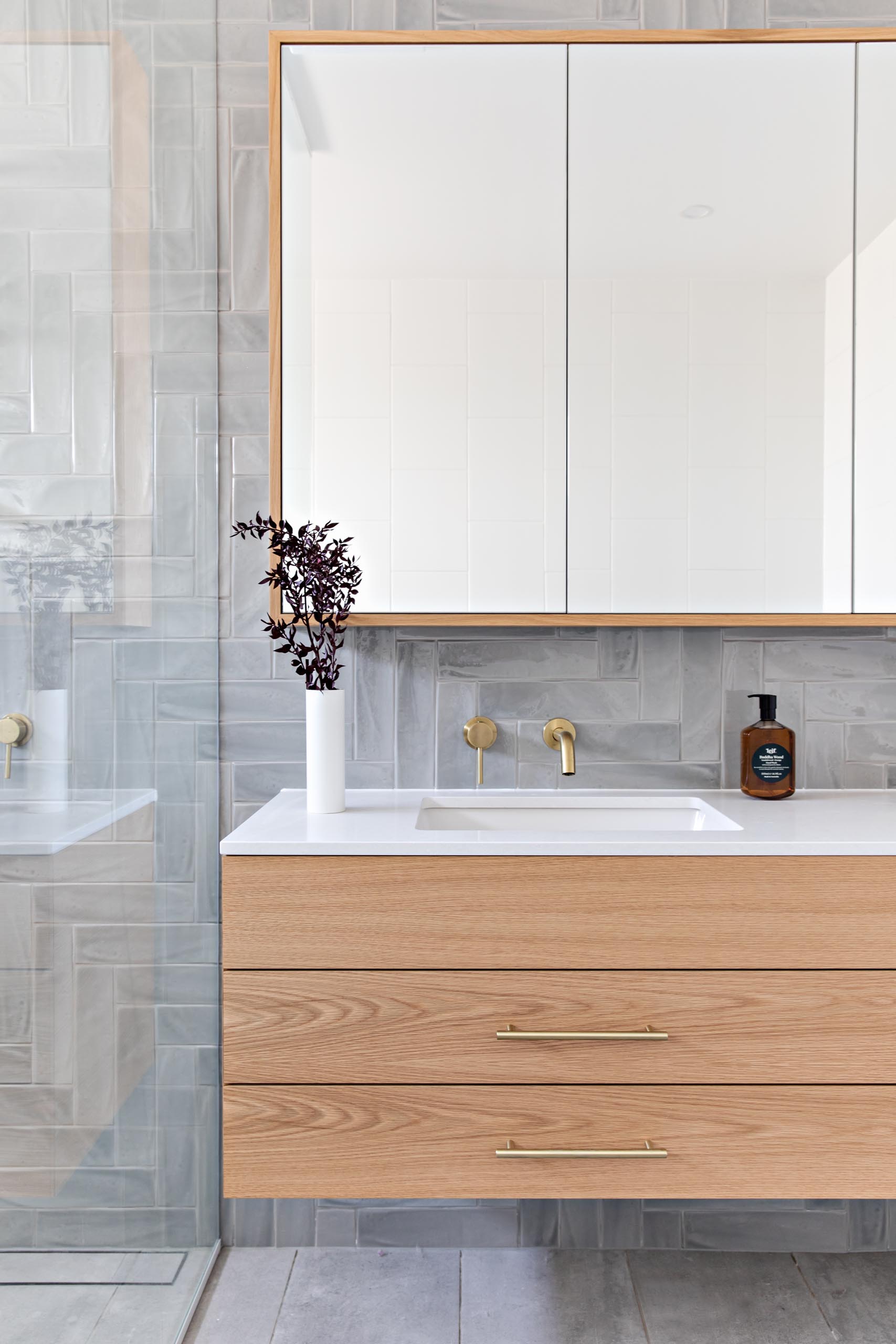A modern bathroom with grey tiles installed in a herringbone pattern cover the wall, a floating wood vanity with a white countertop, and a glass shower screen that separates the shower and freestanding bathtub from the rest of the room.