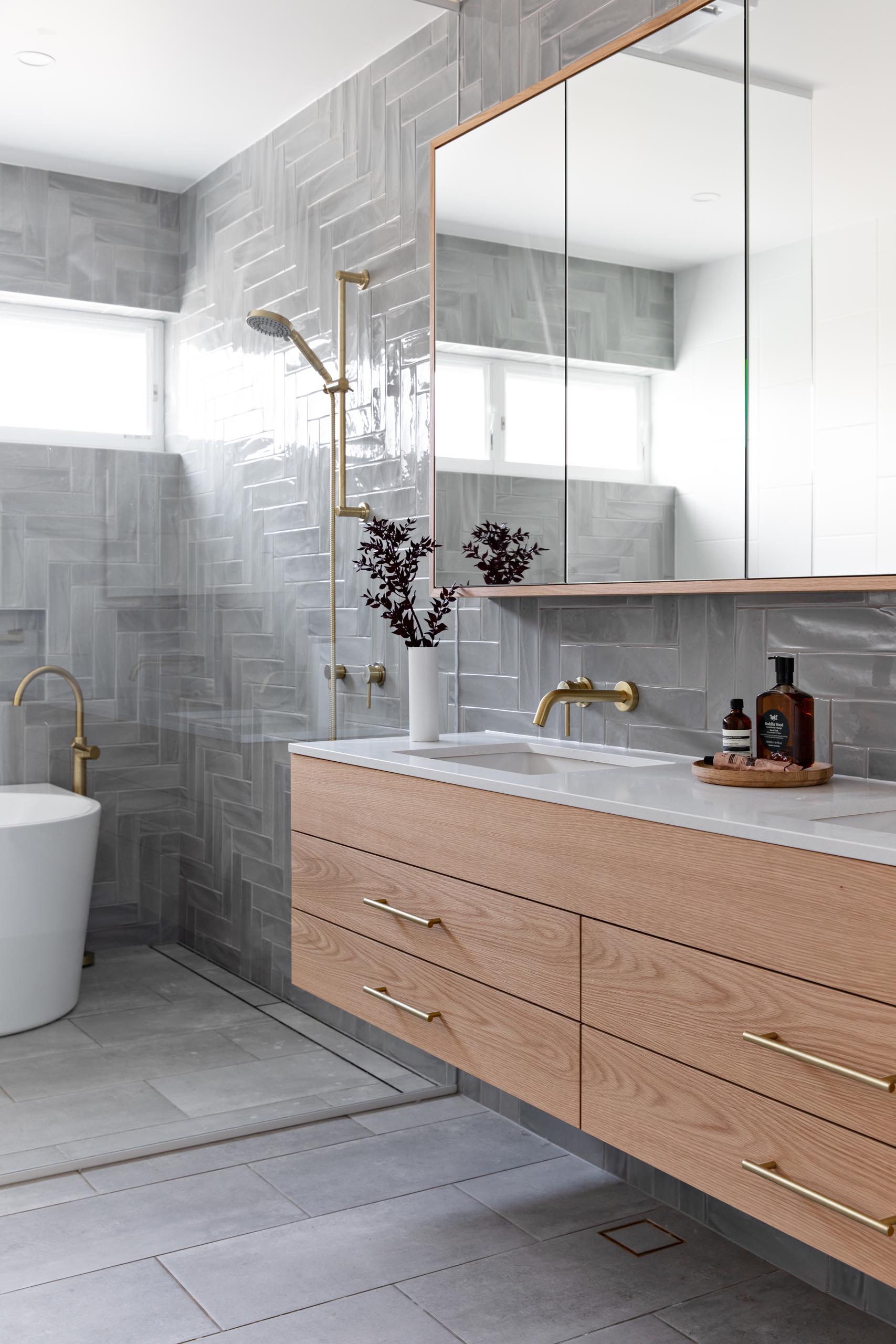 A modern bathroom with grey tiles installed in a herringbone pattern cover the wall, a floating wood vanity with a white countertop, and a glass shower screen that separates the shower and freestanding bathtub from the rest of the room.