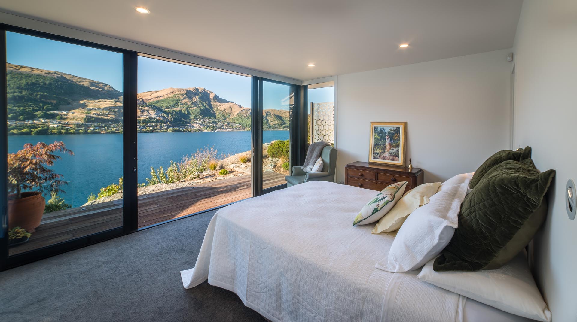 A modern bedroom with floor to ceiling windows and sliding glass doors, that provide an unobstructed view of the lake, which almost looks like a painting.