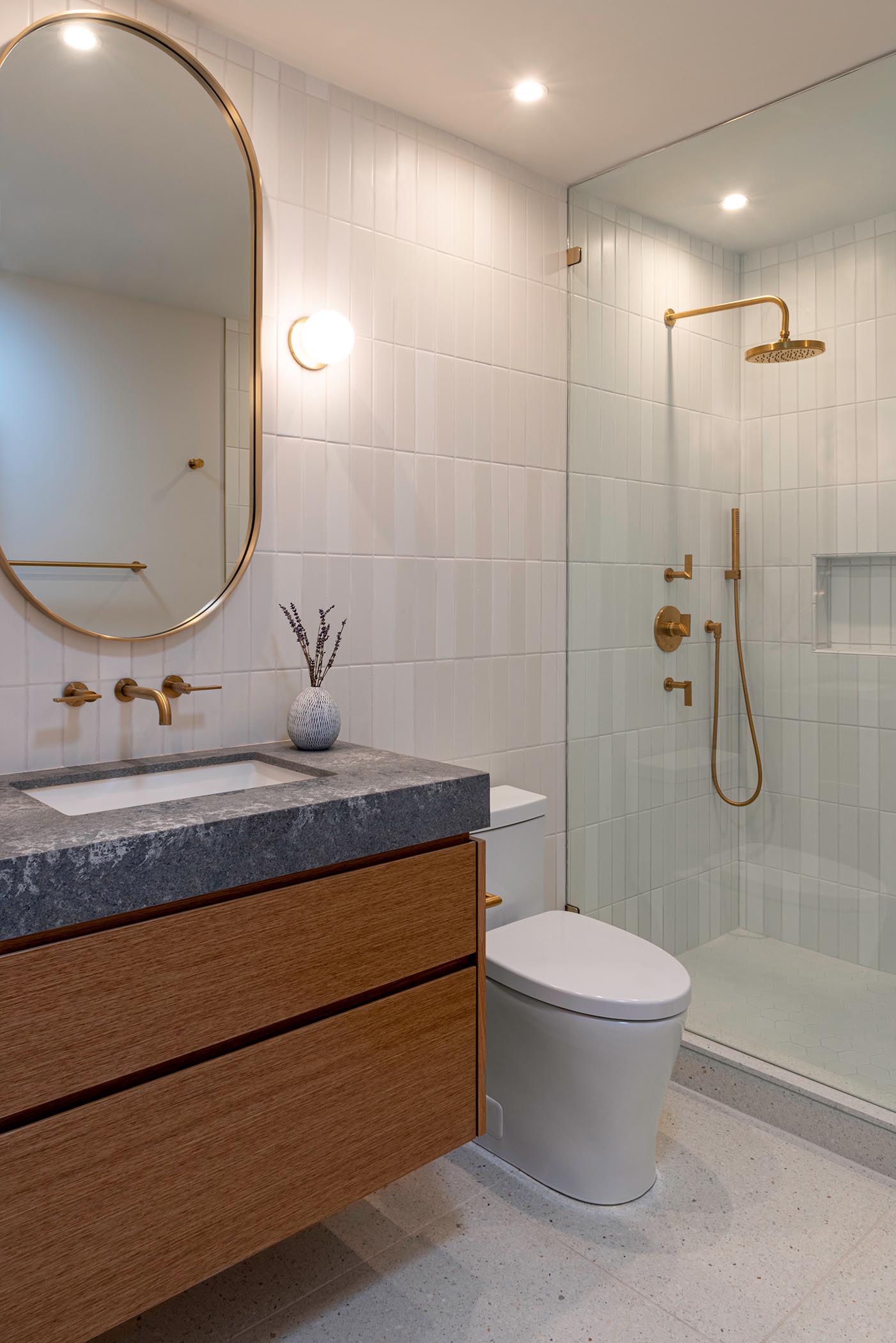 In this bathroom, there's a custom pill-shaped mirror flanked by specialty wall sconces, a walk-in shower with shower niche and a seamless shower screen, white wall tiles, satin brass fixtures, specialty wall sconces, and a Terrazo floor and shower curb.