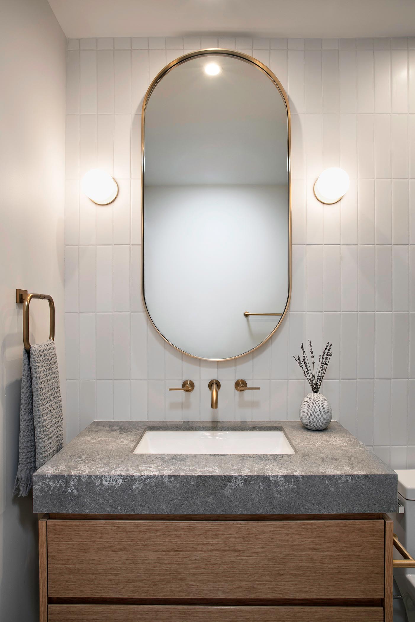 In this bathroom, there's a custom pill-shaped mirror flanked by specialty wall sconces, that hangs above a wood vanity with a grey countertop and white undermount sink.