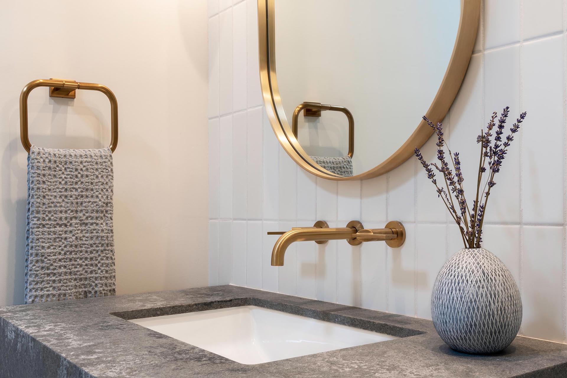 In this bathroom, there's a custom pill-shaped mirror flanked by specialty wall sconces, that hangs above a wood vanity with a grey countertop and white undermount sink.