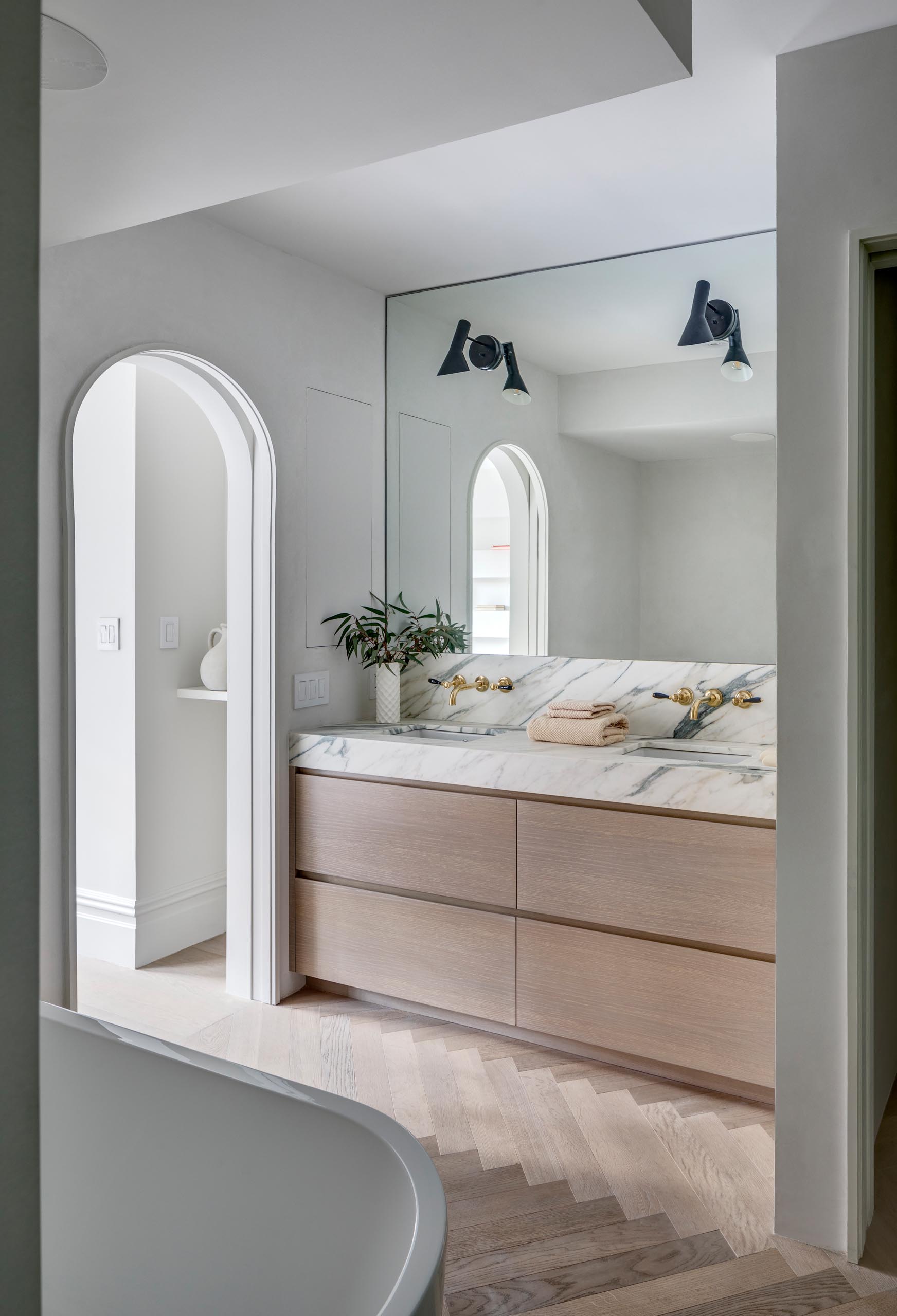 A modern master bathroom with an arched doorway, a wood and marble vanity, and a wood floor laid in a herringbone pattern.