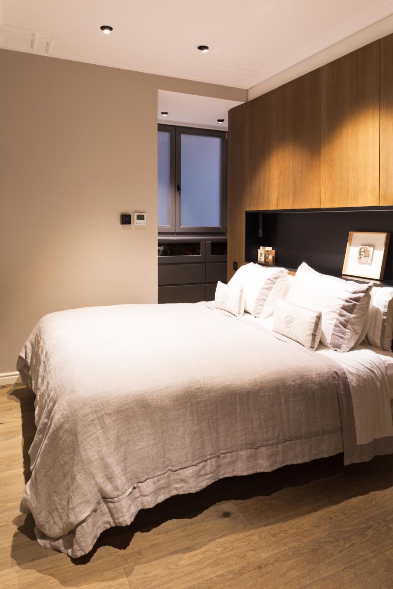 In this modern master bedroom, there's a wood accent wall behind the bed that includes a matte black shelving niche with lighting.