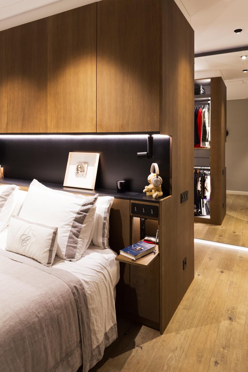 In this modern master bedroom, there's a wood accent wall behind the bed that includes a matte black shelving niche with lighting.