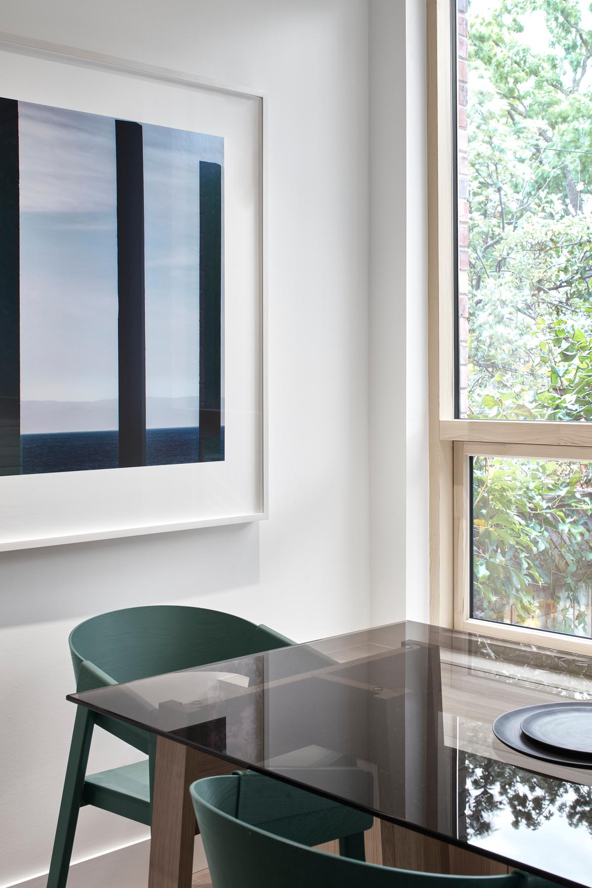 A modern dining room with a glass topped dining table, green chairs, artwork, and views of the backyard.