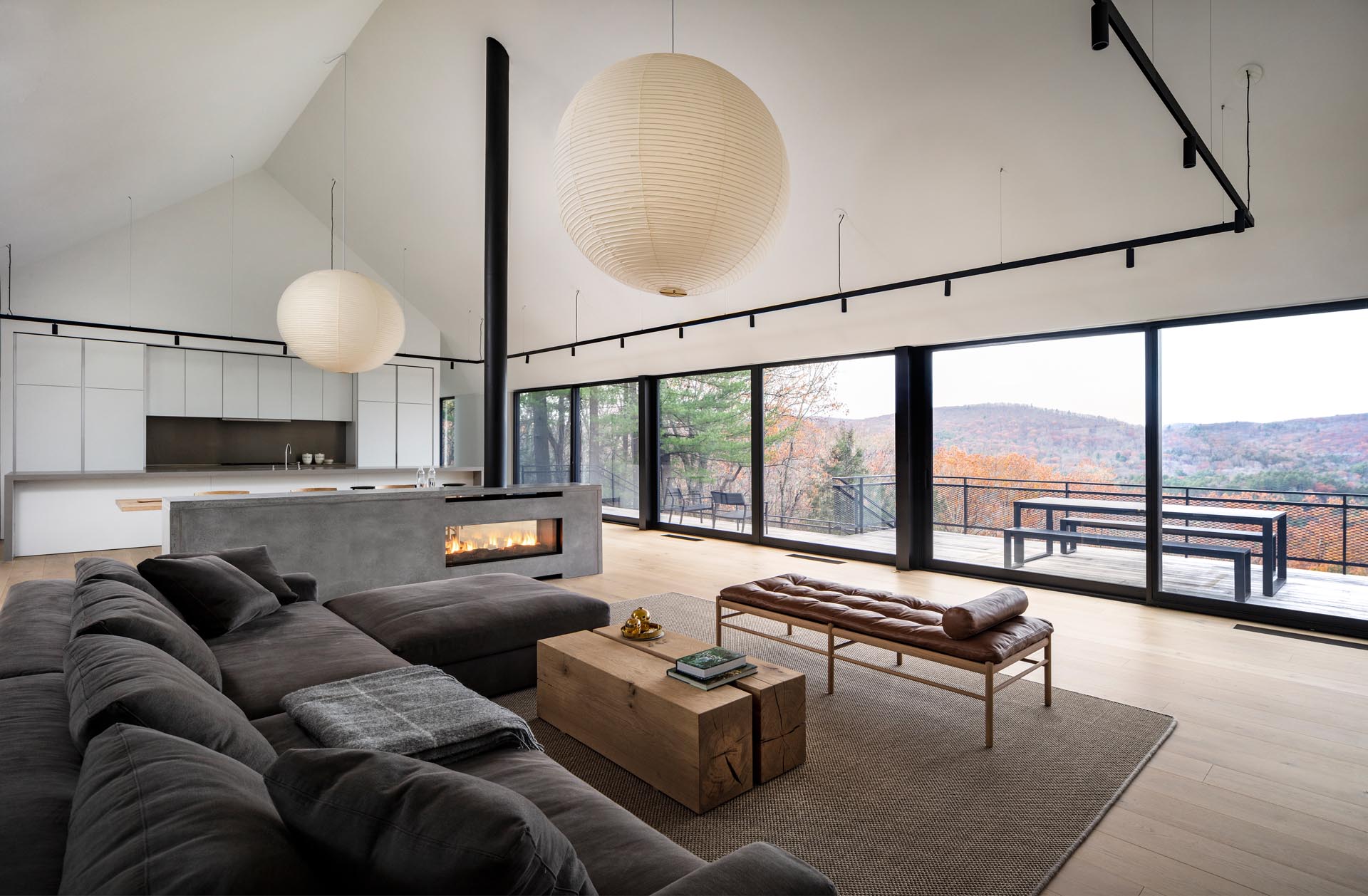 A modern house interior with a great room that includes the living room, a concrete fireplace, the dining room, and a white kitchen.