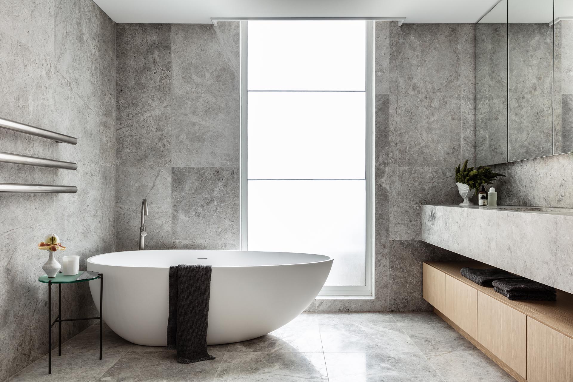 In this modern master bathroom, natural stone with a grey finish covers the walls and floors, as well as the vanity. A floor-to-ceiling frosted window allows the light to pass through, but still allows for privacy.