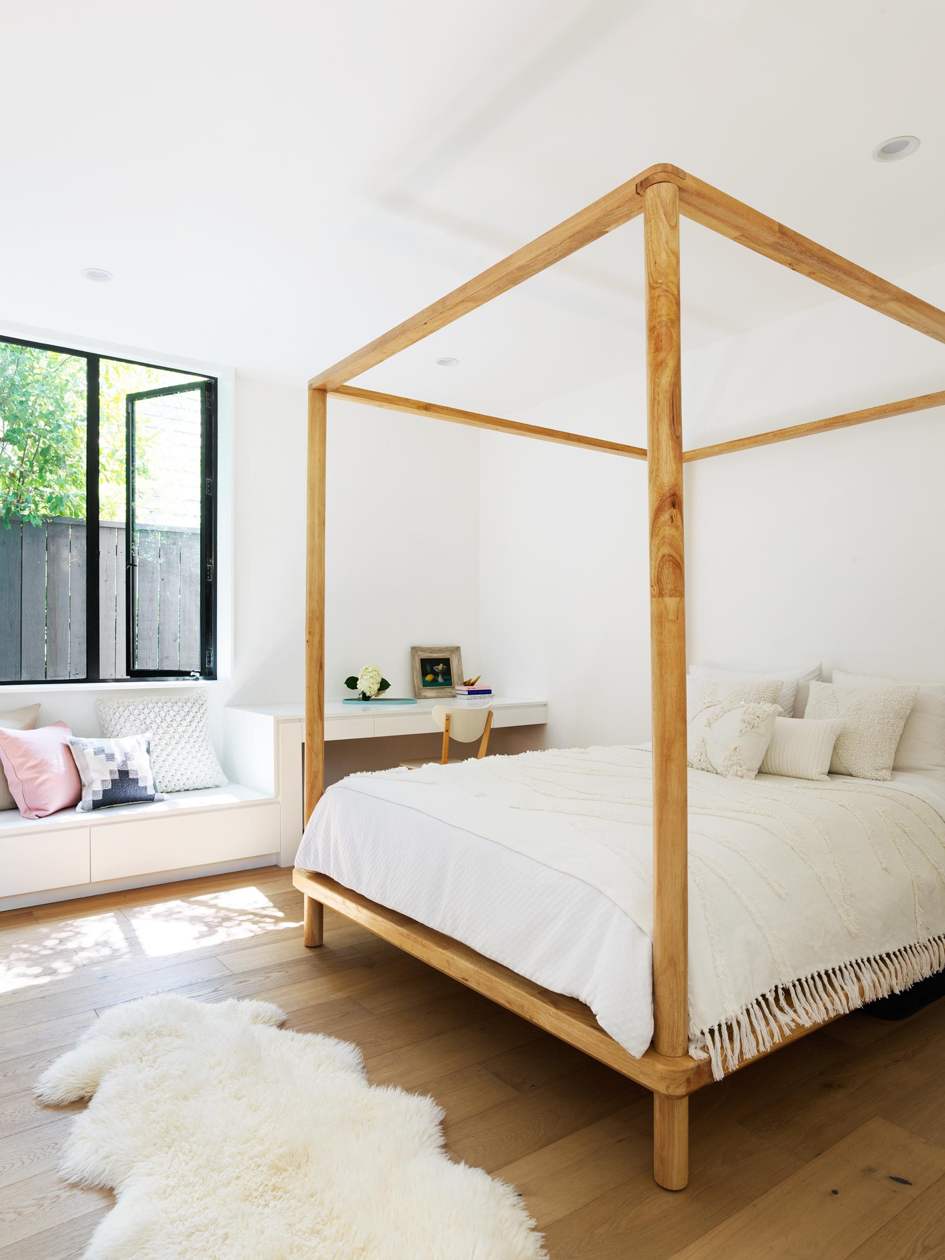 A kids bedroom with a four poster wood bed frame, and along the wall is a built-in desk and bench.