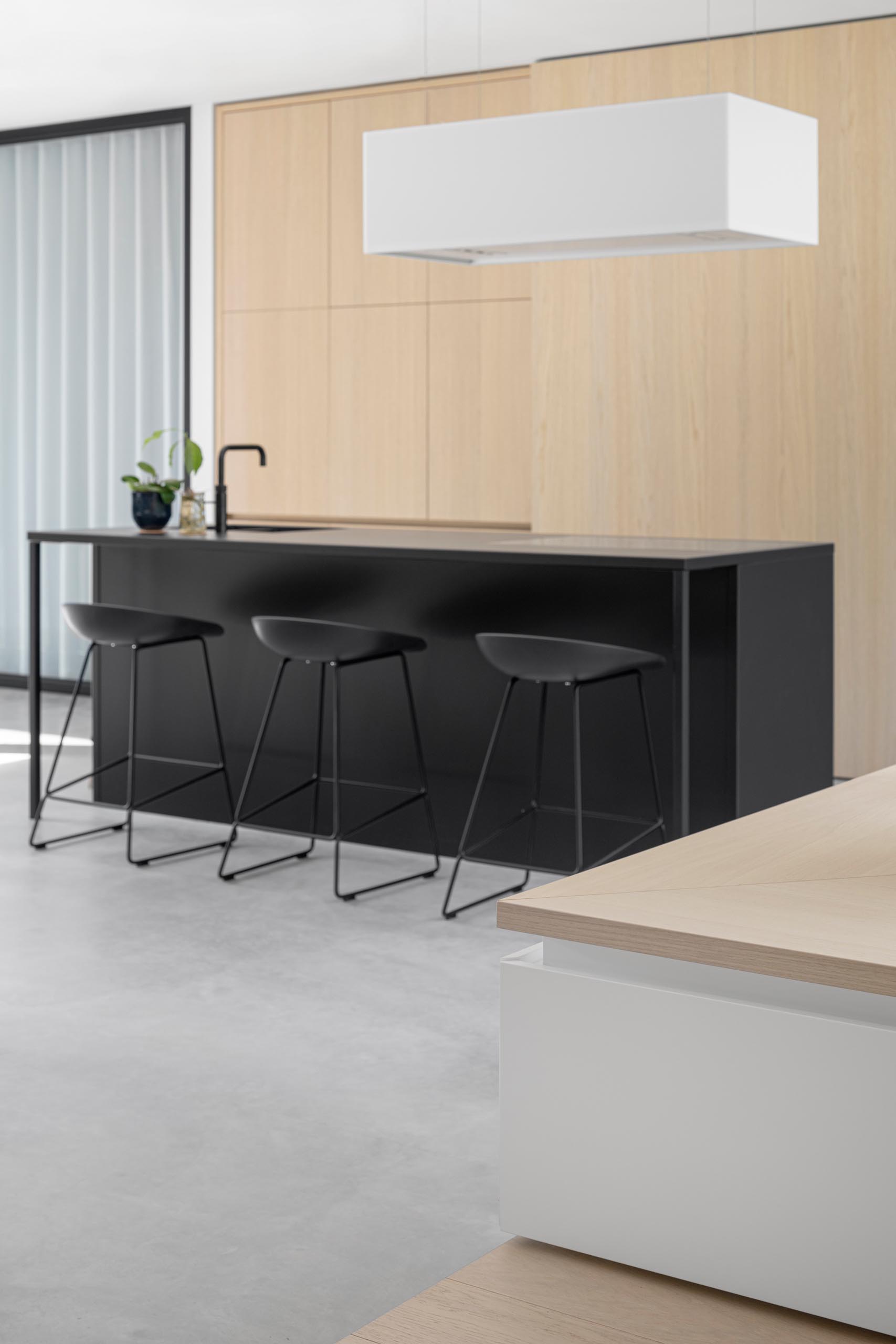 A modern kitchen with minimalist wood cabinets, a matte black island with matching stools, and concrete floors.