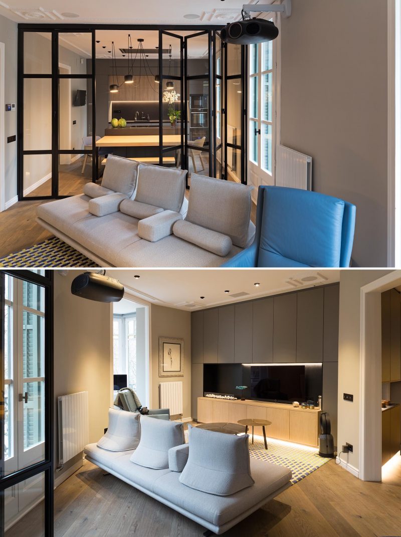 A modern living room in a remodeled apartment has a wall of cabinetry, and a black-framed glass wall that separates it from the kitchen / dining room.