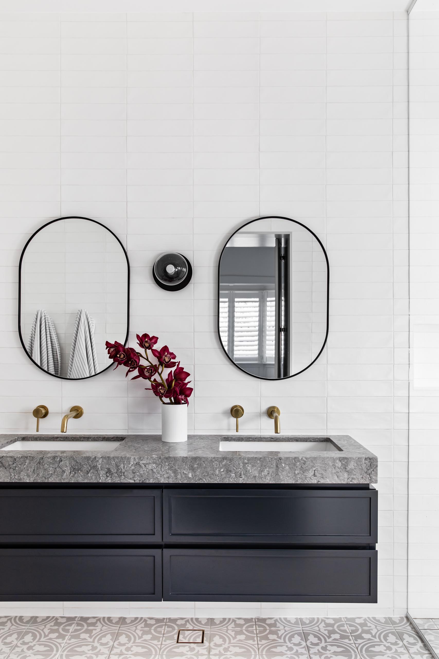 A modern bathroom with patterned floor tiles, a floating black vanity topped with grey stone, white subway tiles that cover the walls, oval black-framed mirrors, and brass hardware