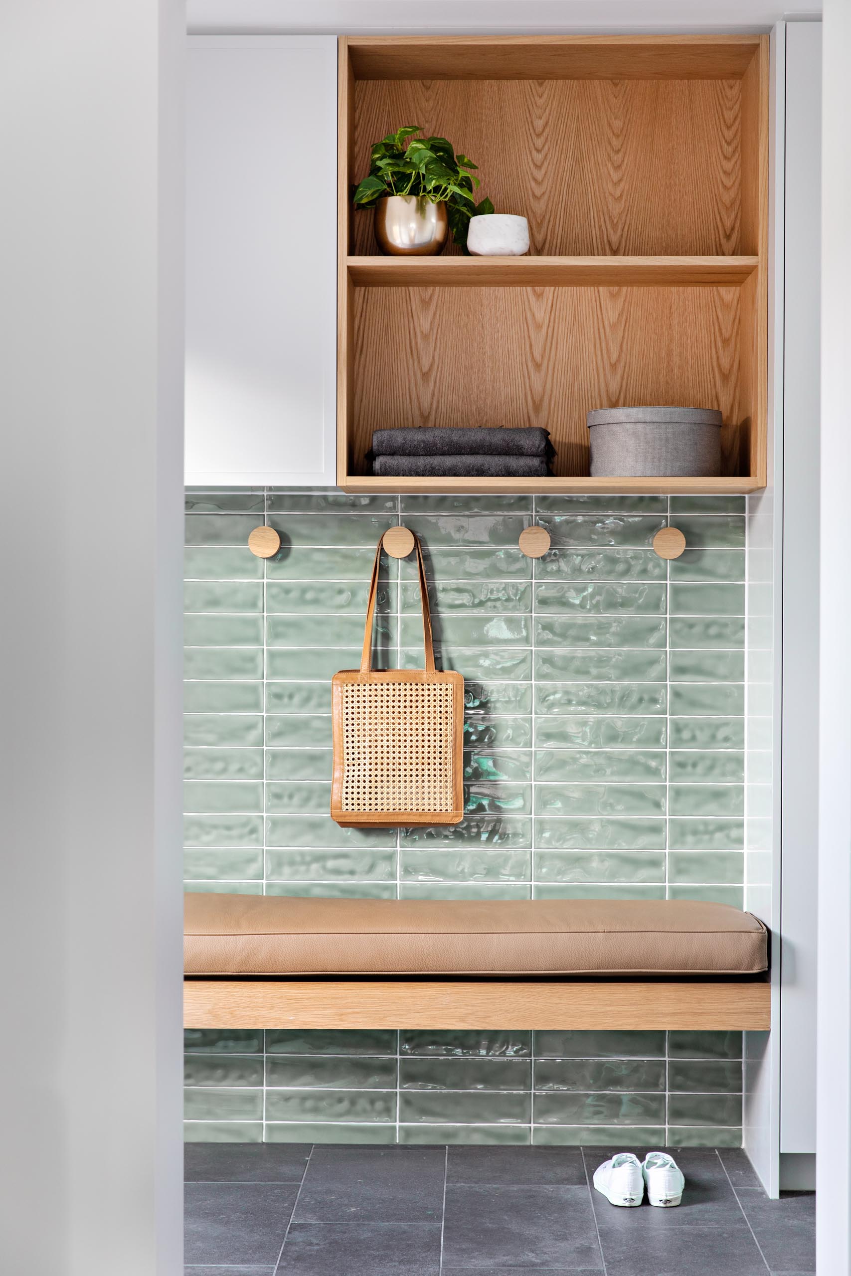 A small mud room with soft green tiles, a wood bench with brown leather cushion, round wood hooks, and a cabinet with open wood shelving.