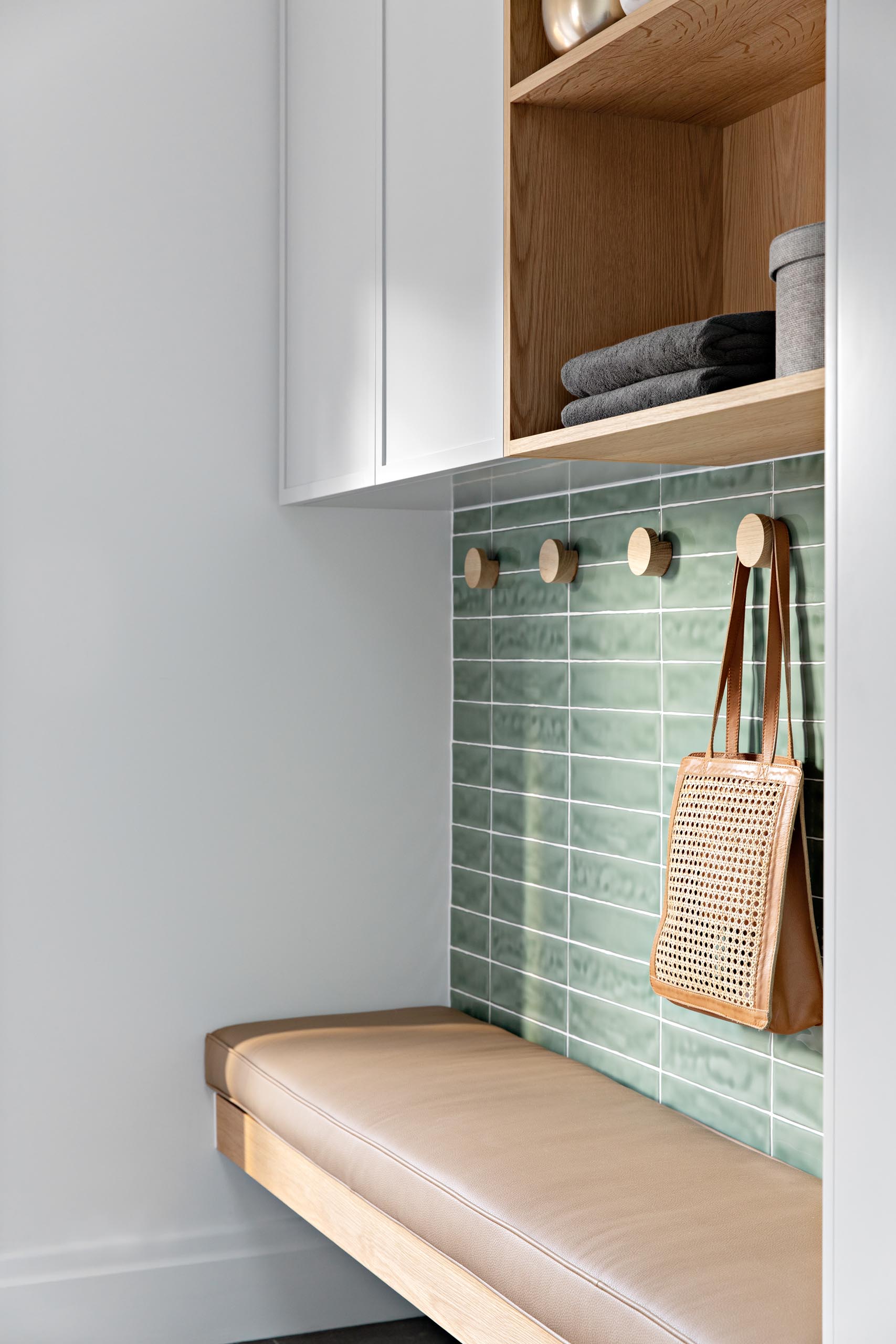 A small mud room with soft green tiles, a wood bench with brown leather cushion, round wood hooks, and a cabinet with open wood shelving.