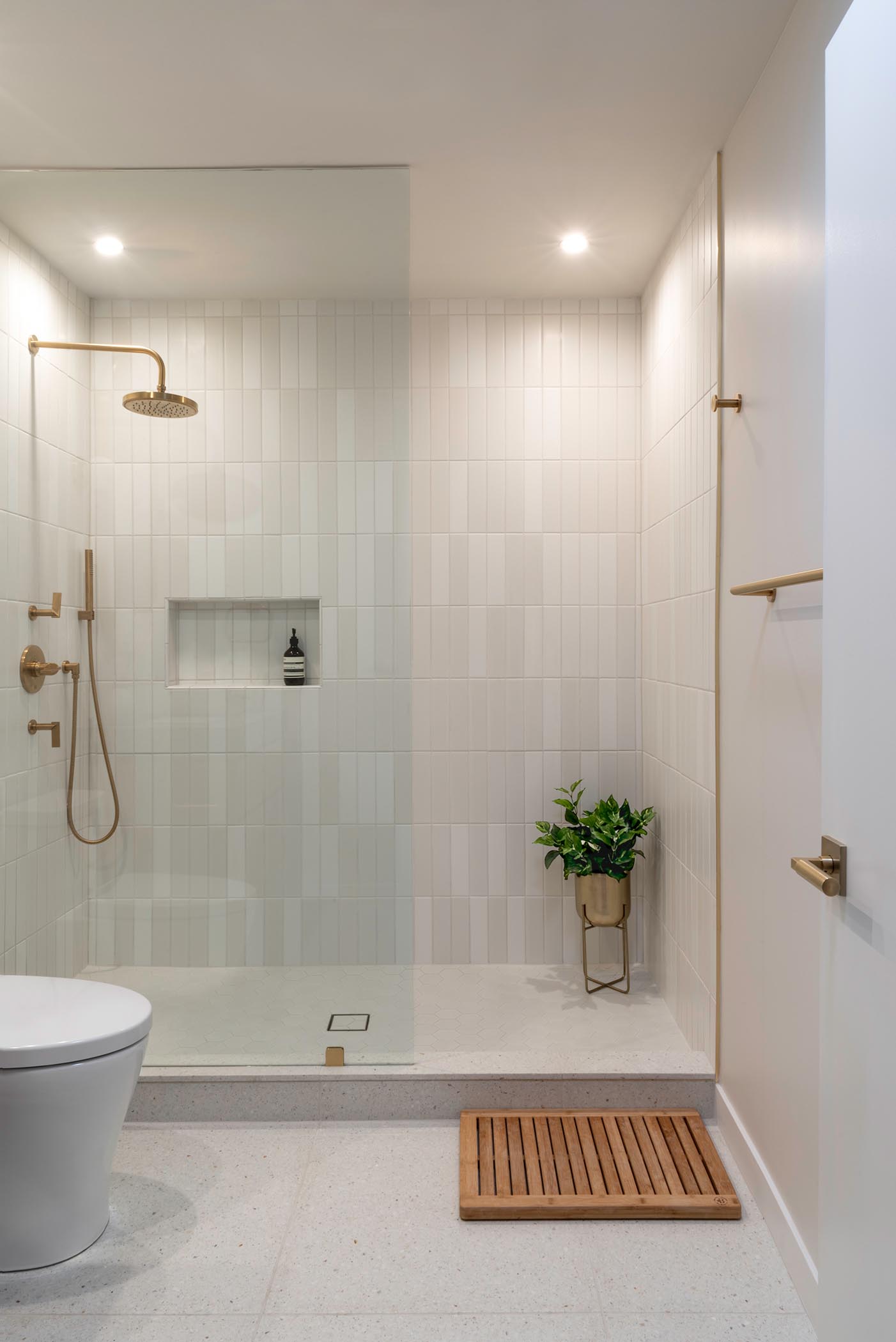 In this bathroom, there's a walk-in shower with shower niche and a seamless shower screen, white wall tiles, satin brass fixtures, and  Terrazo floor and shower curb.