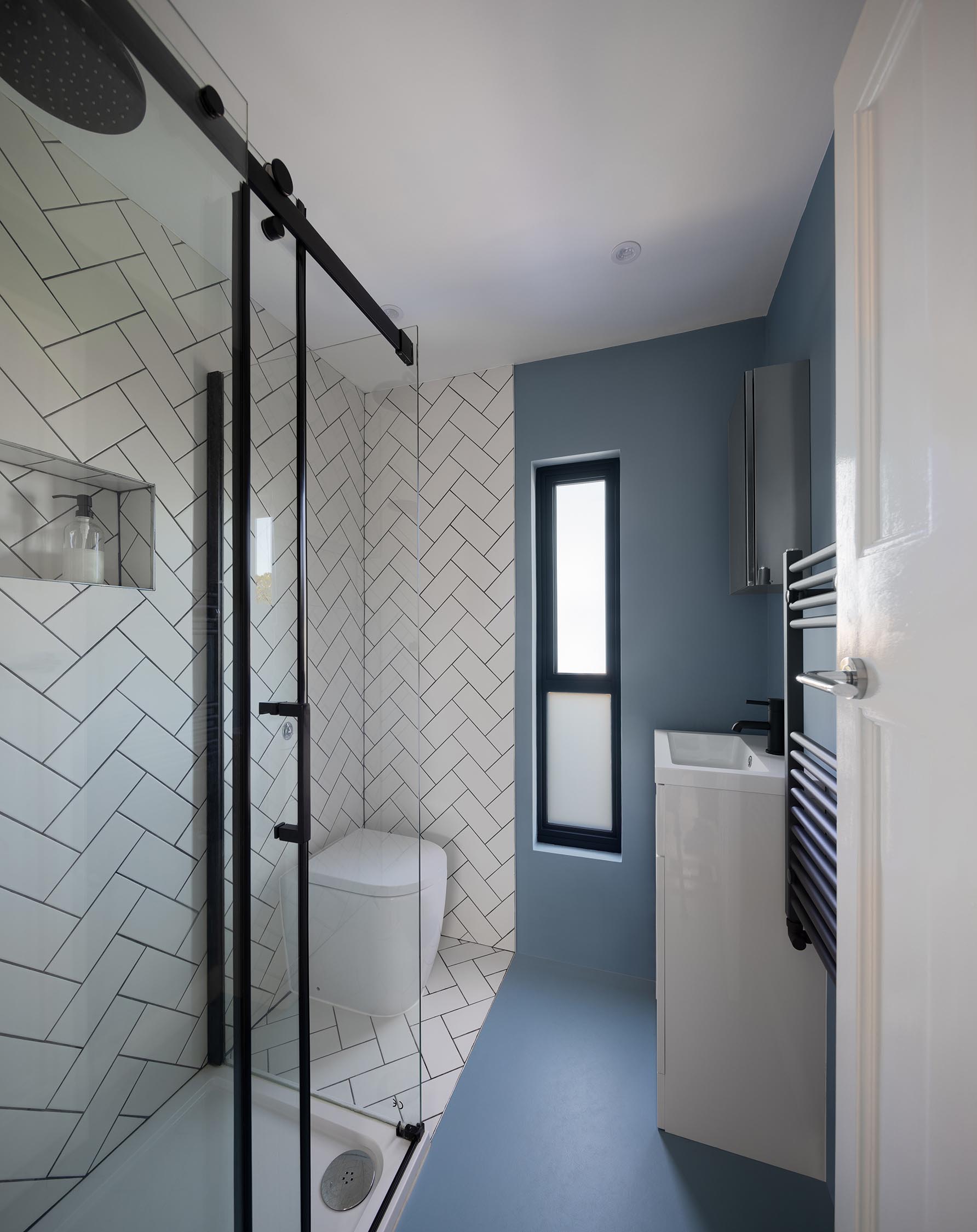 A modern bathroom with white tiles in a herringbone pattern, black hardware, and narrow vanity.